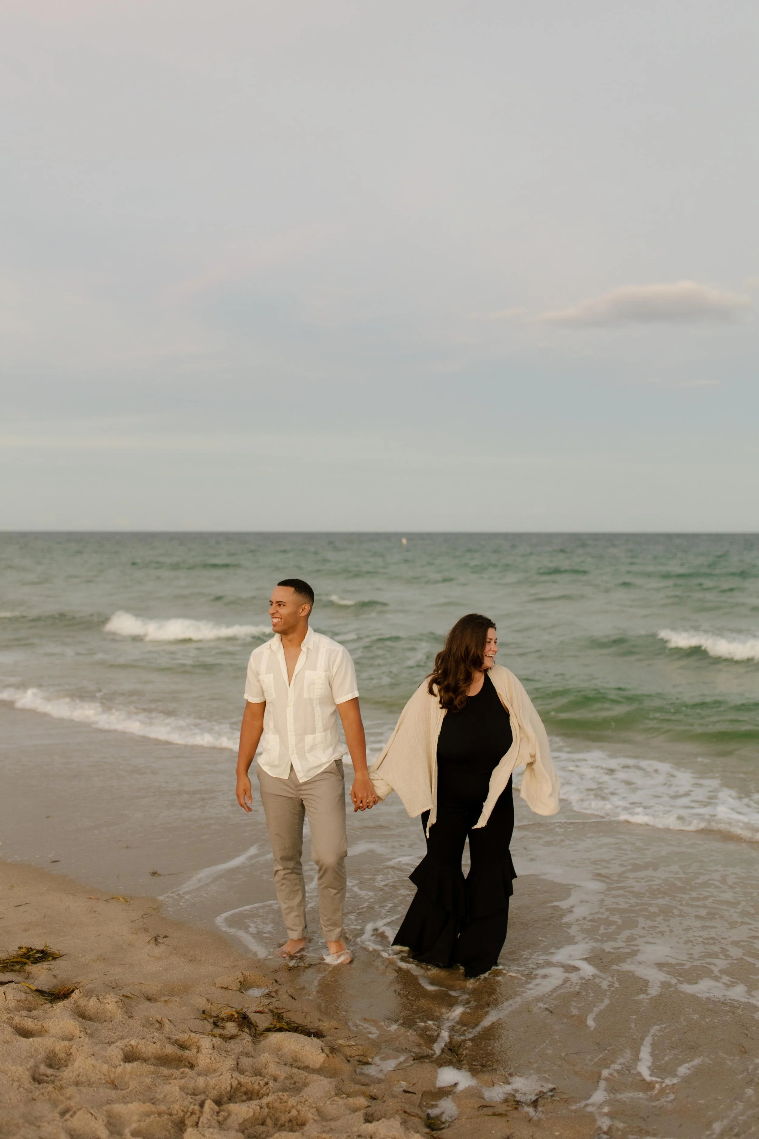 Couple walking barefoot along the shoreline holding hands, waves washing over their feet as they look off in opposite directions, grounded and peaceful