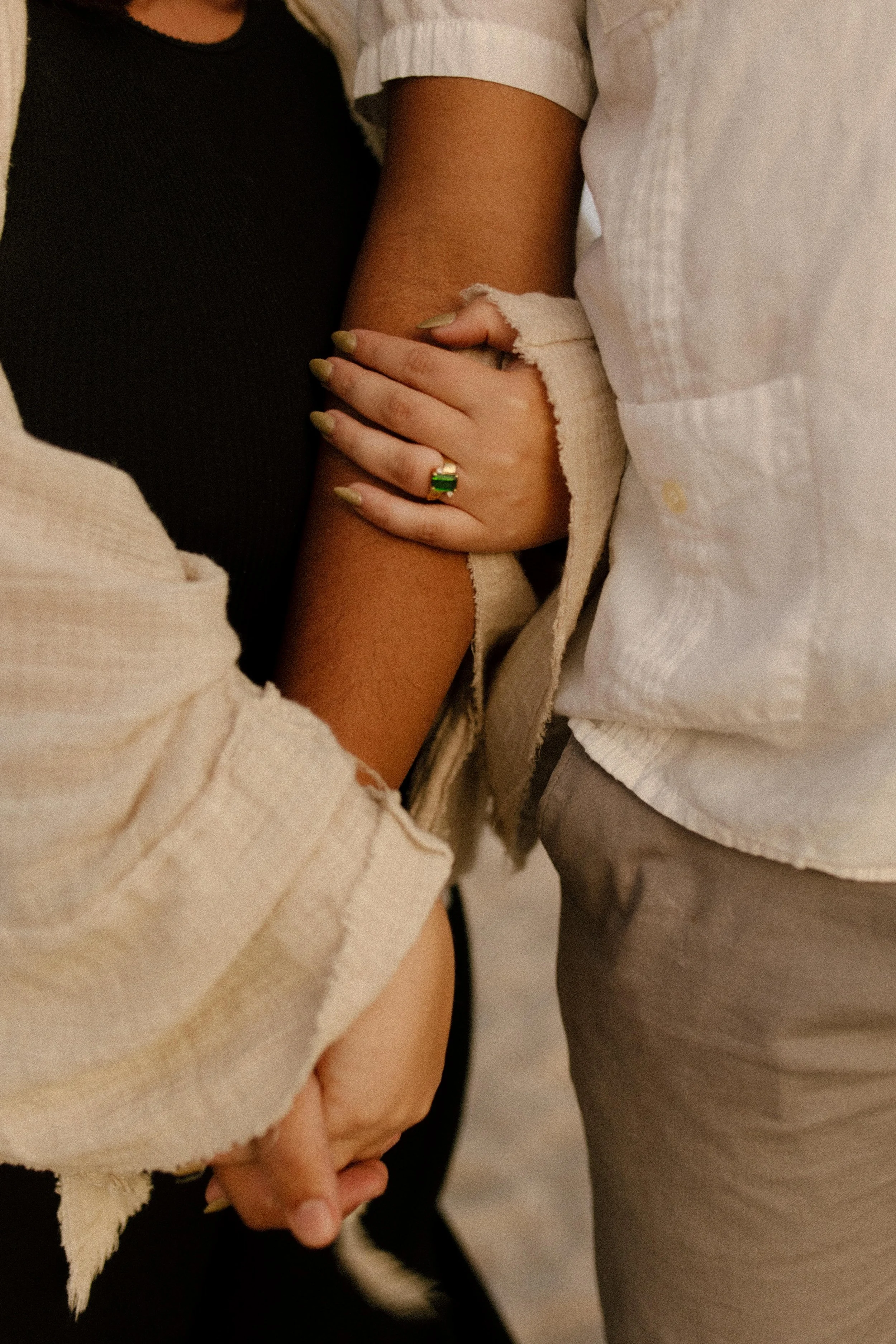 Close-up of hands intertwined, highlighting a green gemstone engagement ring against soft, neutral tones and golden light
