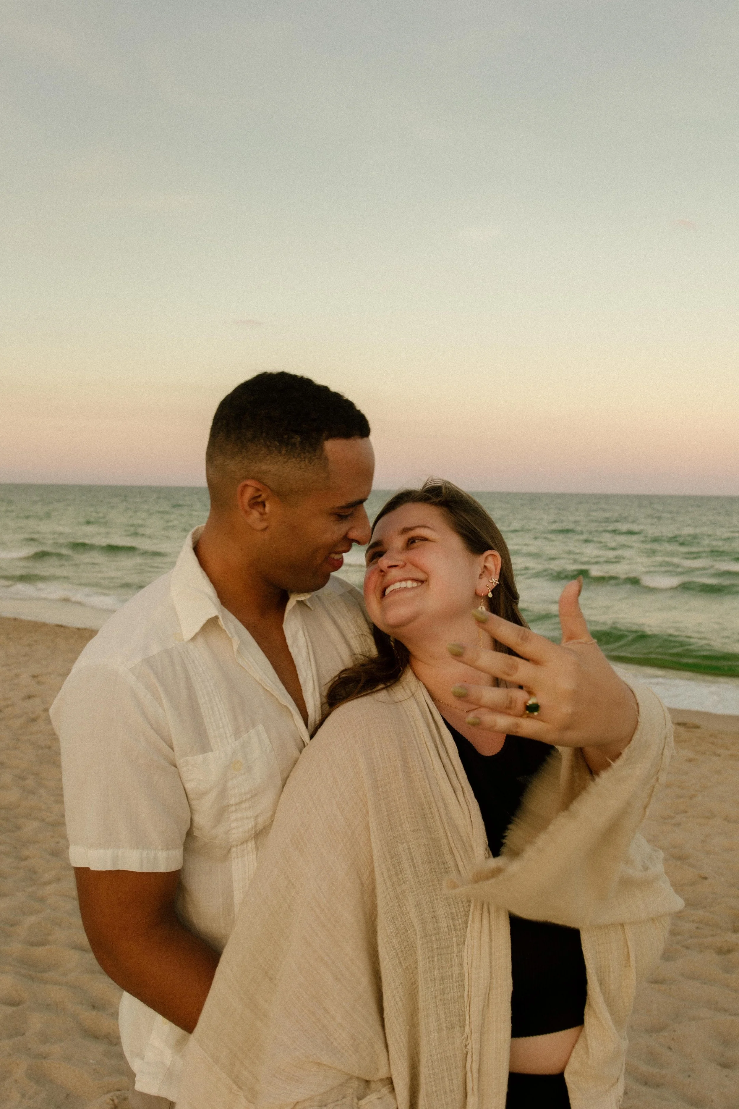 Man and woman embracing on the beach at sunset, her hand lifted to show a new engagement ring as they smile into each other, golden light wrapping around them