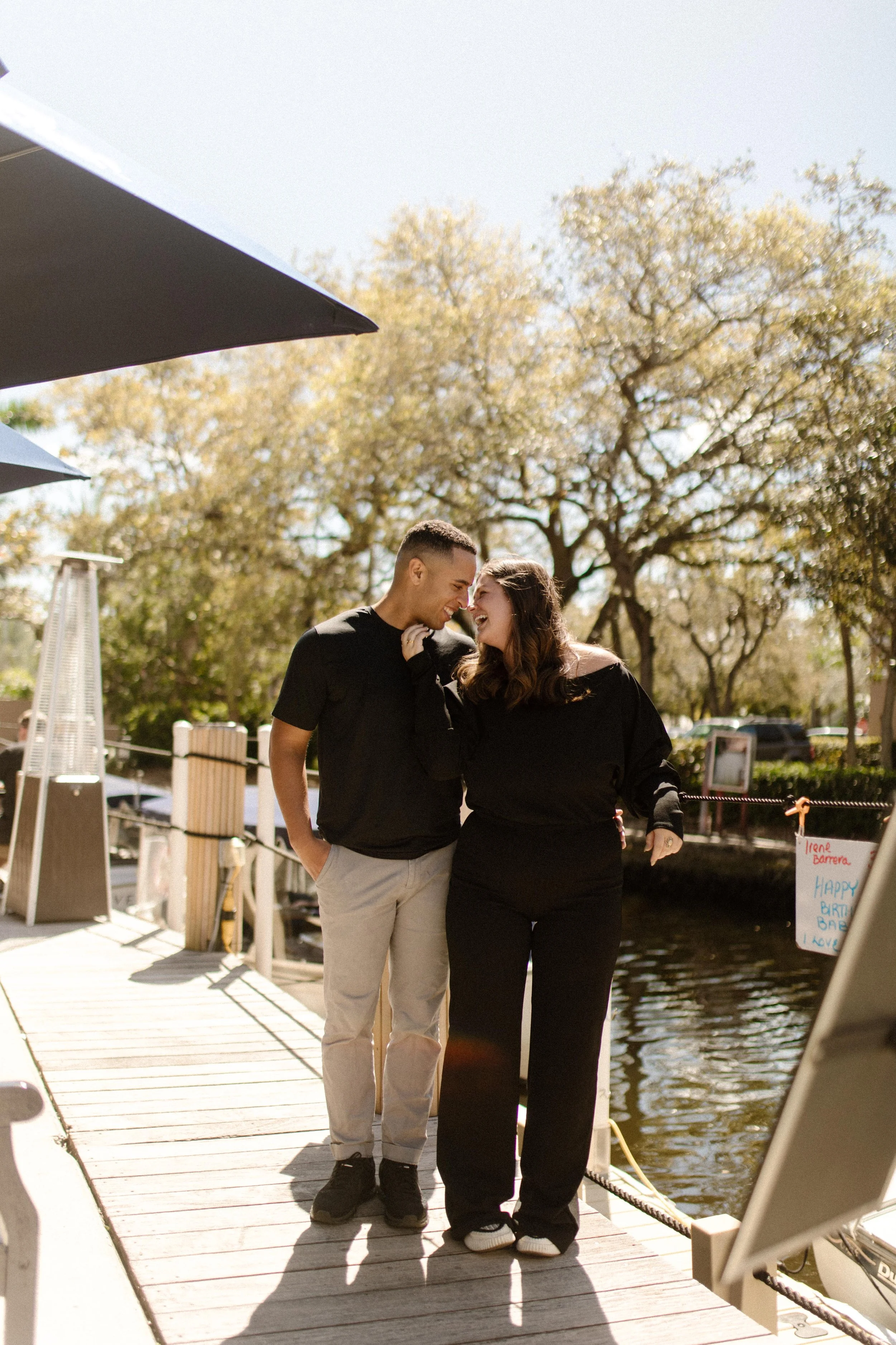Couple walking closely together on a sunny dock, laughing and leaning into each other beside the water, capturing the kind of quiet lead-up that makes you think about how to plan a surprise proposal