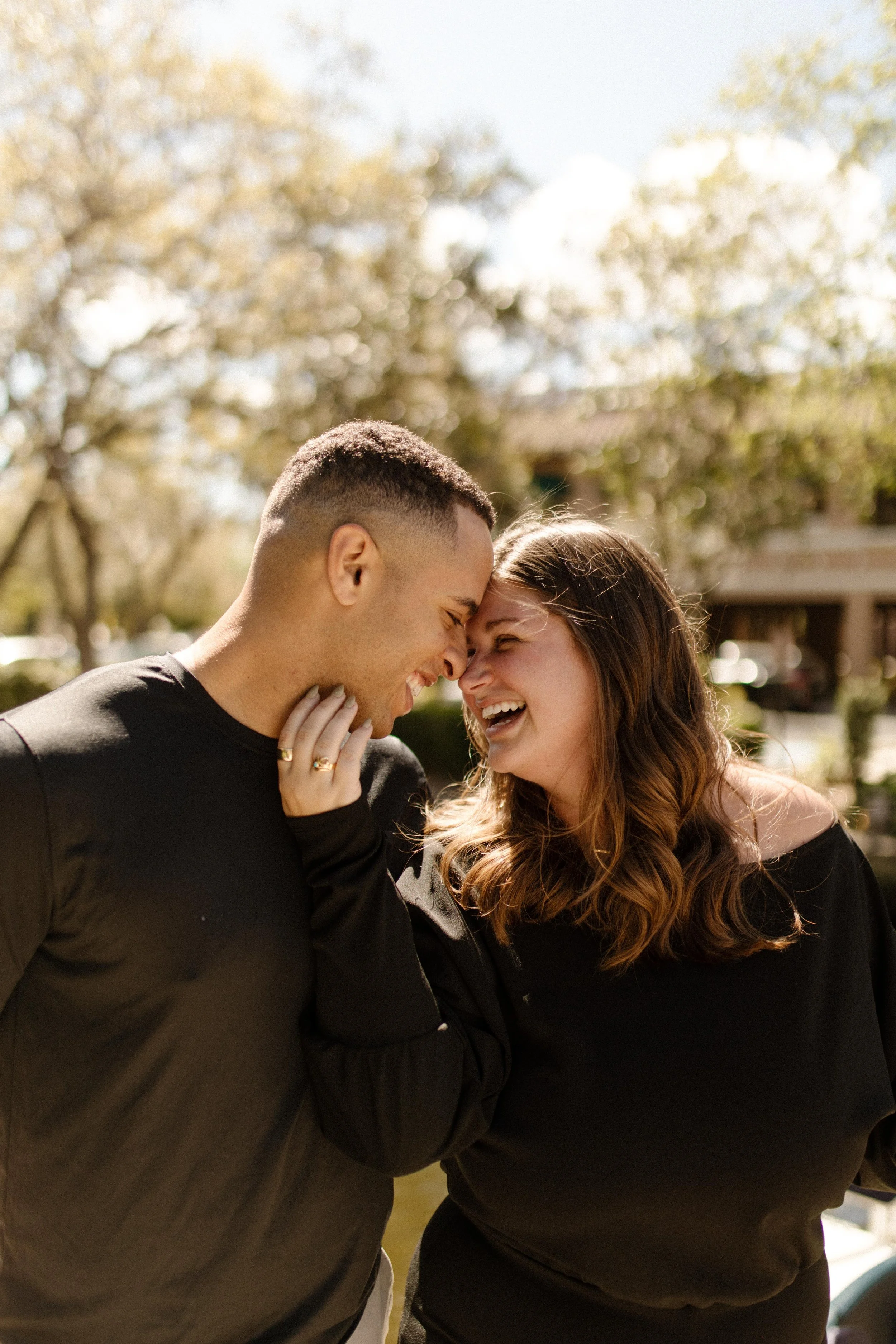 Close-up of a couple laughing with their foreheads pressed together in warm sunlight, her hand resting on his neck, completely lost in each other