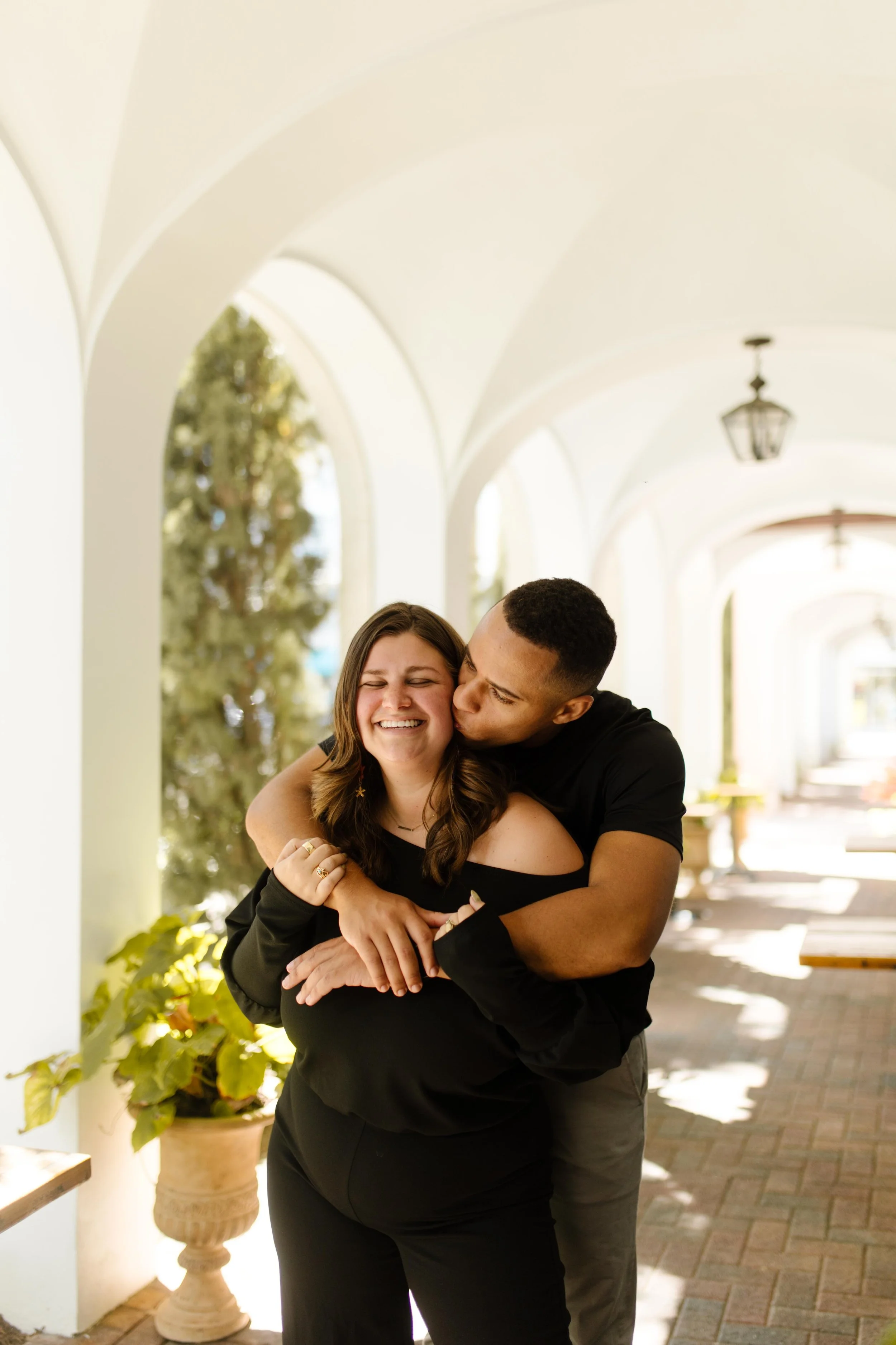 Man wrapping his arms around his partner under a sunlit archway, kissing her cheek while she smiles with her eyes closed