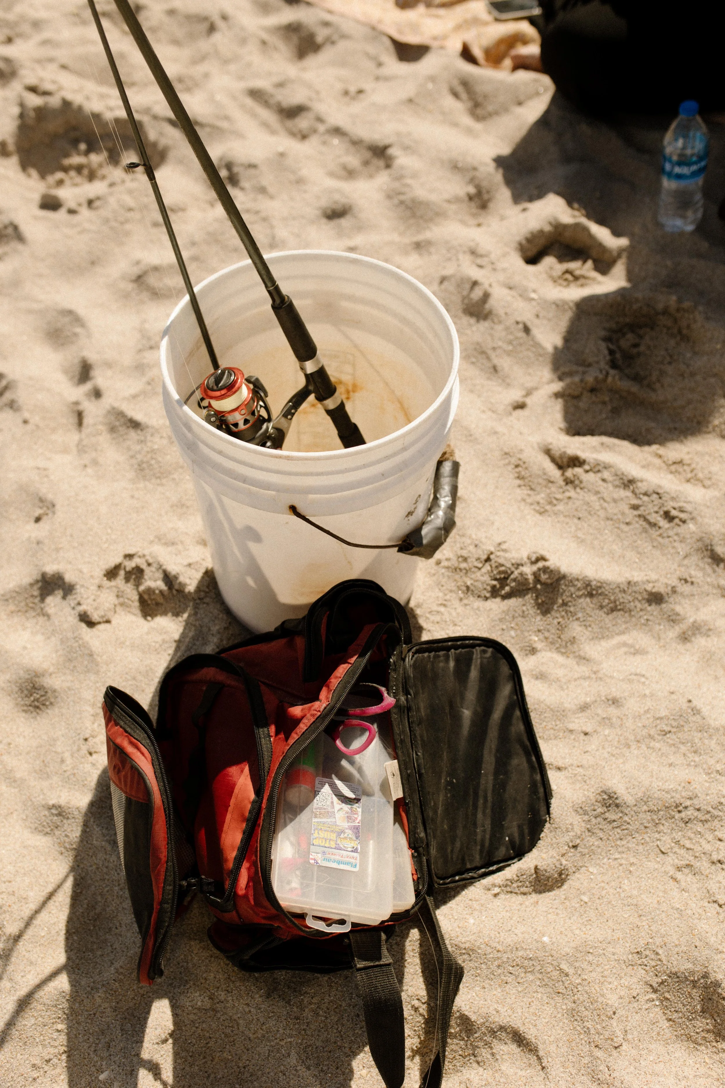 Fishing gear in a white bucket and an open tackle bag resting on the sand, a subtle detail that plays into how to plan a surprise proposal with a believable cover story