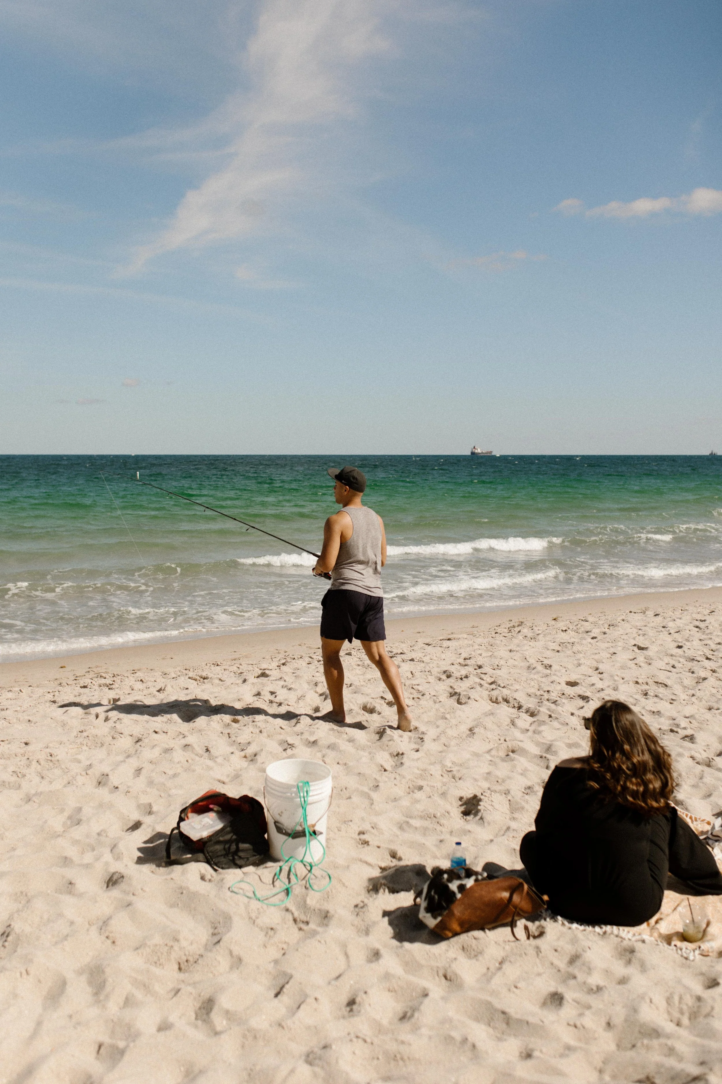 Man walking toward the shoreline with a fishing rod while a woman sits on a blanket behind him, watching the waves roll in