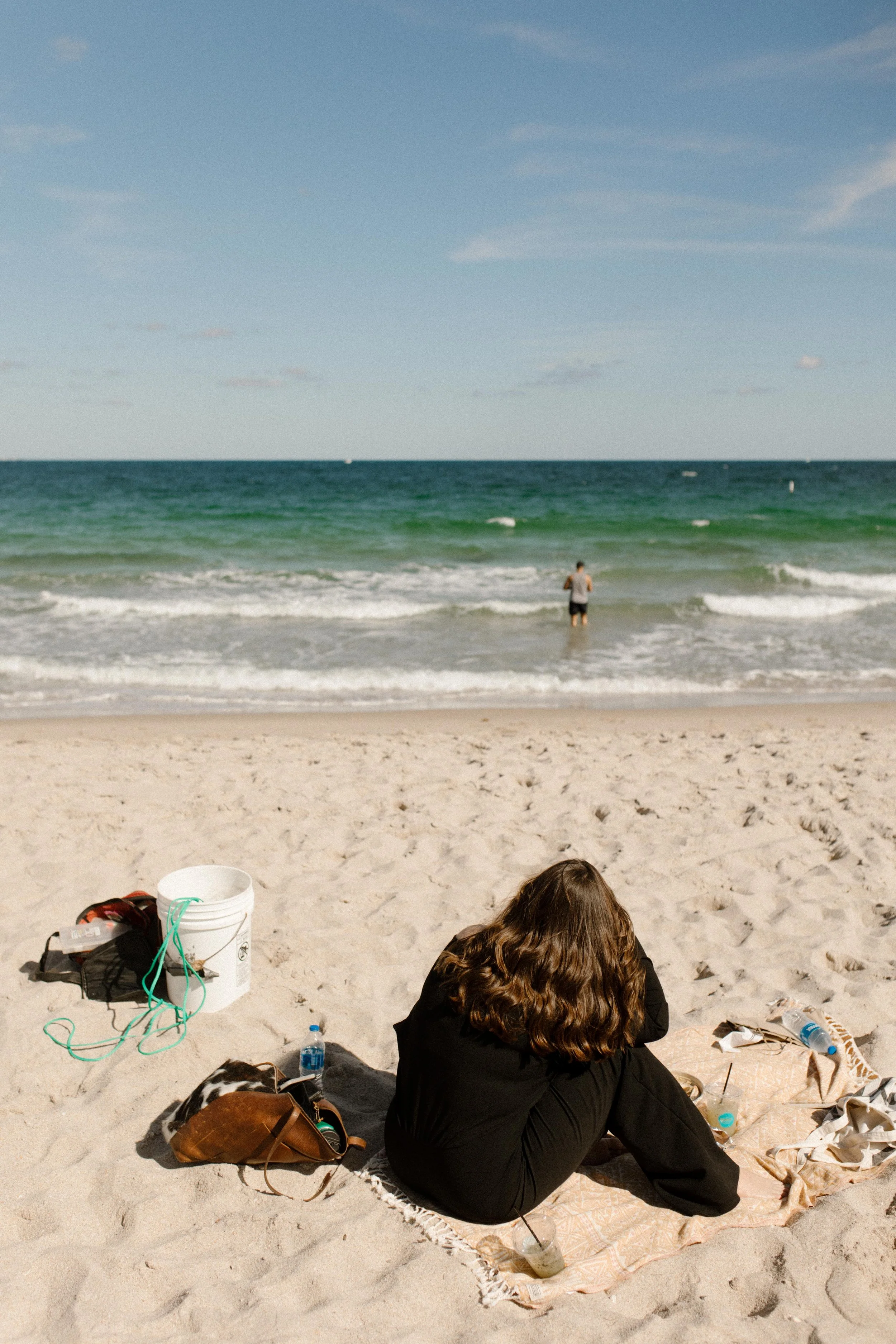 Woman sitting on a beach blanket facing the ocean, surrounded by personal belongings and soft afternoon light, a quiet pause that feels like how to plan a surprise proposal