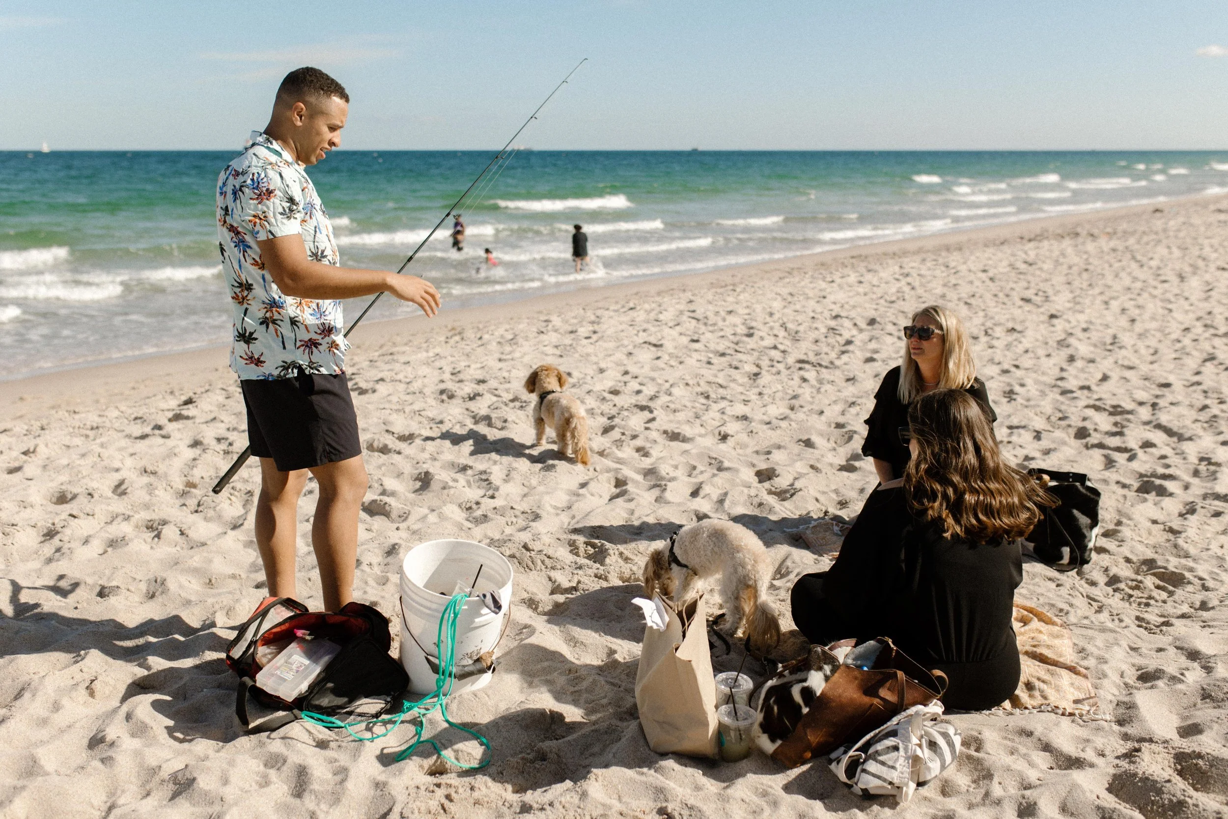 Man holding a fishing rod on the beach while two women sit nearby with dogs and bags, a casual scene that quietly mirrors how to plan a surprise proposal without raising suspicion
