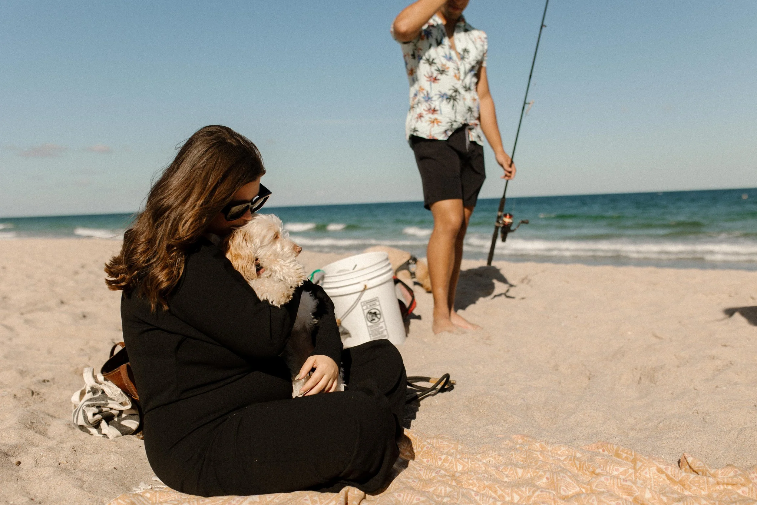 Woman sitting on a beach blanket holding a small dog while a man stands nearby with a fishing rod, a relaxed and unsuspecting moment that mirrors how to plan a surprise proposal