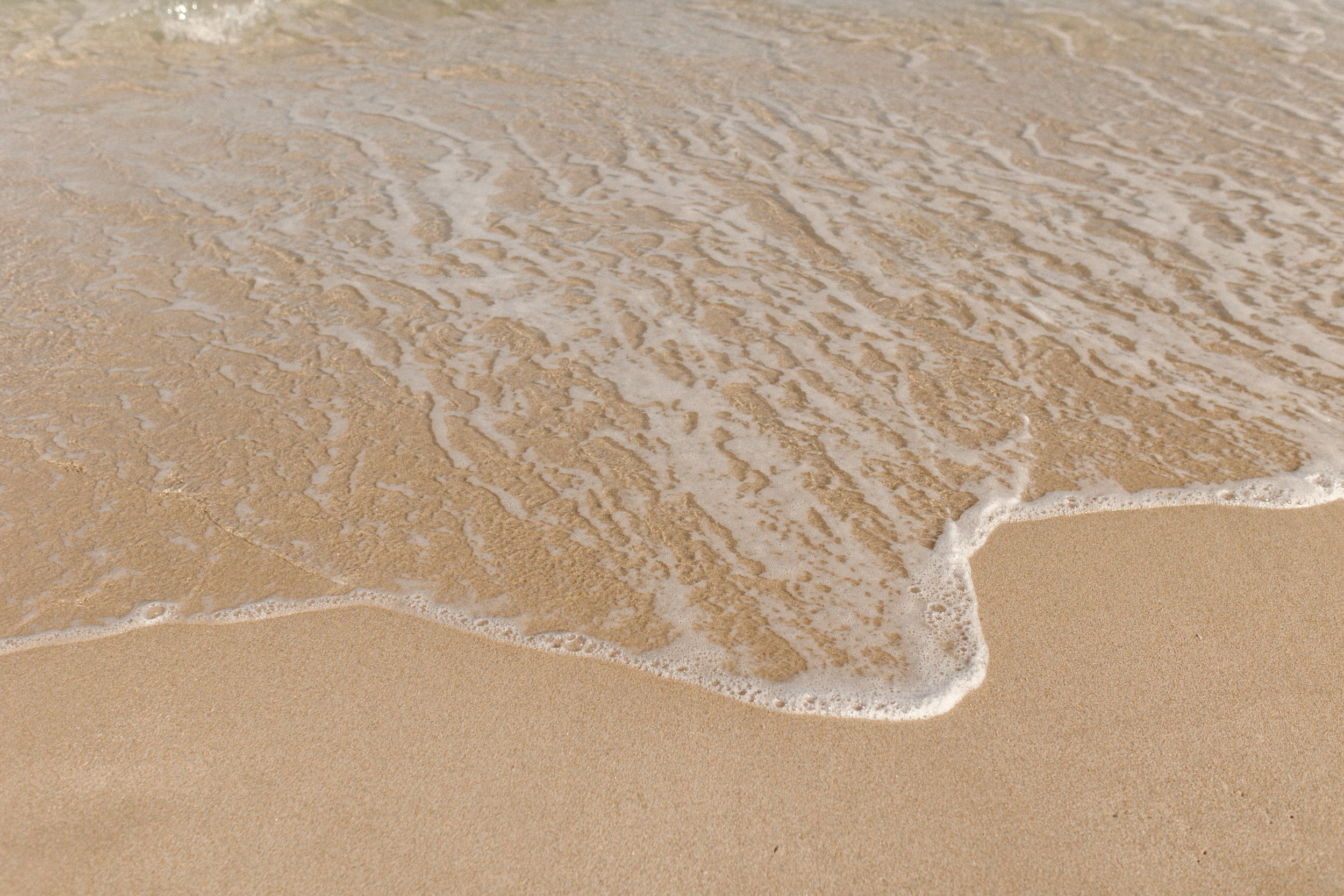 Close-up of ocean water washing over textured sand, soft foam tracing the shoreline in a peaceful, grounding moment between memories