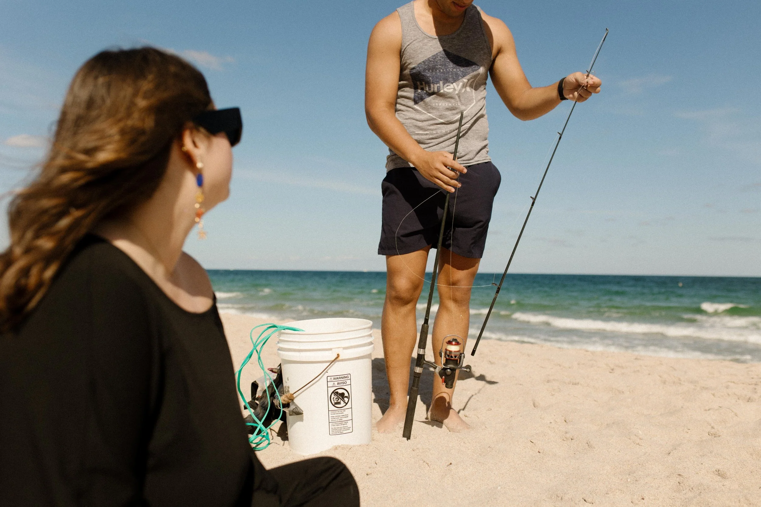 Man holding fishing gear while standing in the sand as a woman watches from a blanket nearby, a believable cover that plays into how to plan a surprise proposal
