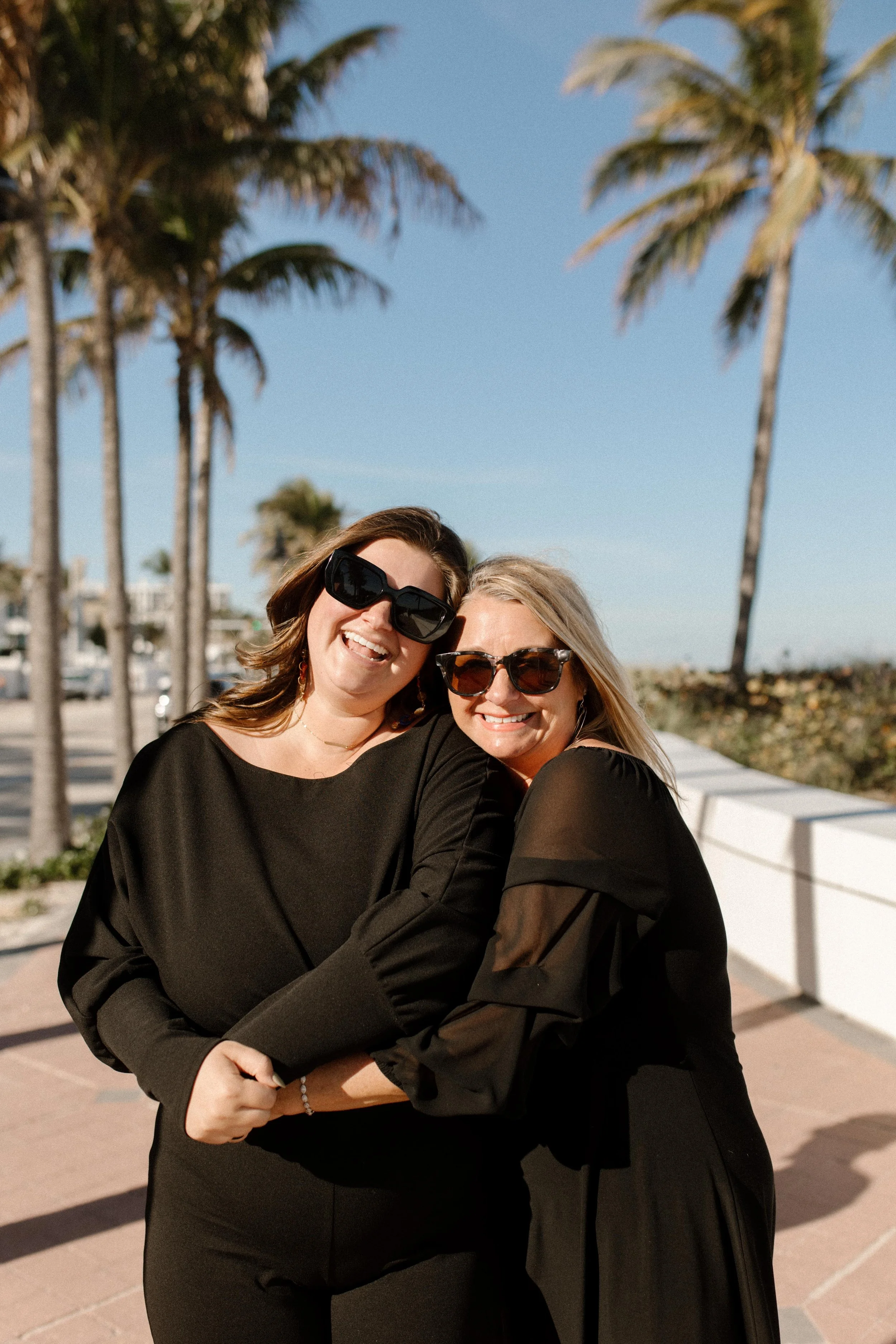 Two women hugging and smiling on a sunny sidewalk lined with palm trees, joy radiating in a way that feels like celebrating how to plan a surprise proposal gone right