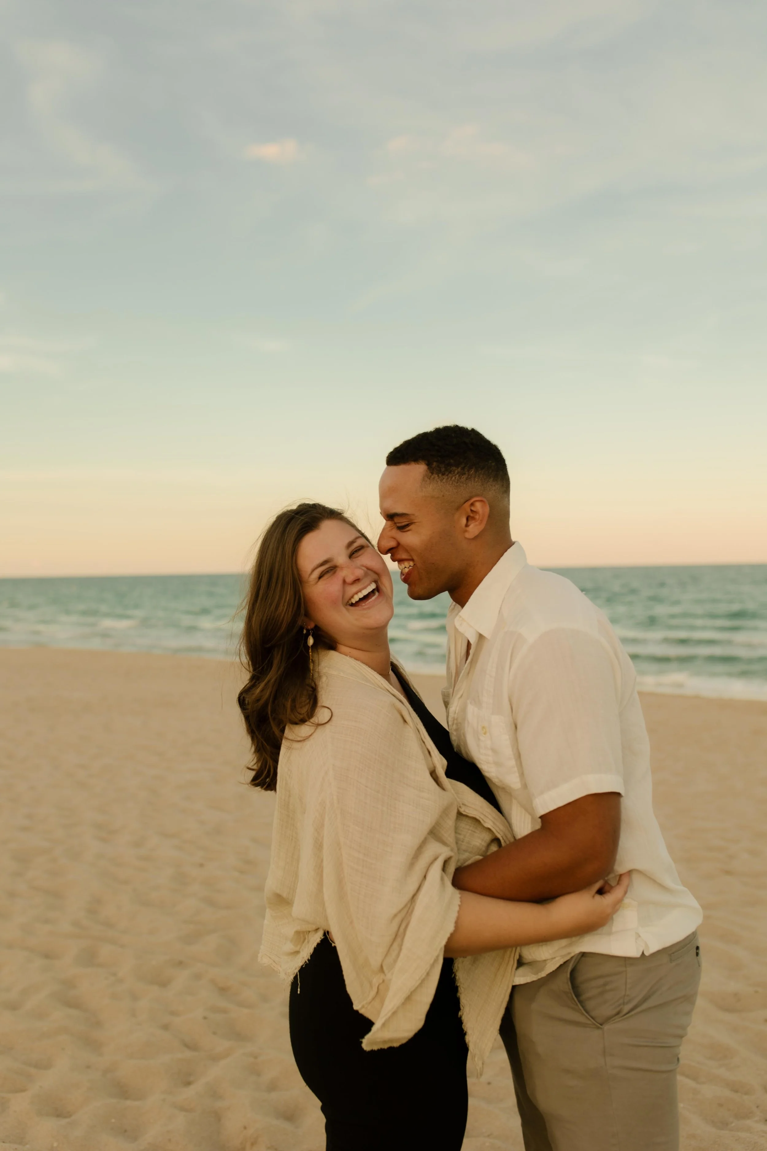 Couple laughing together on the beach at sunset, bodies turned toward each other with wind in her hair and waves behind them