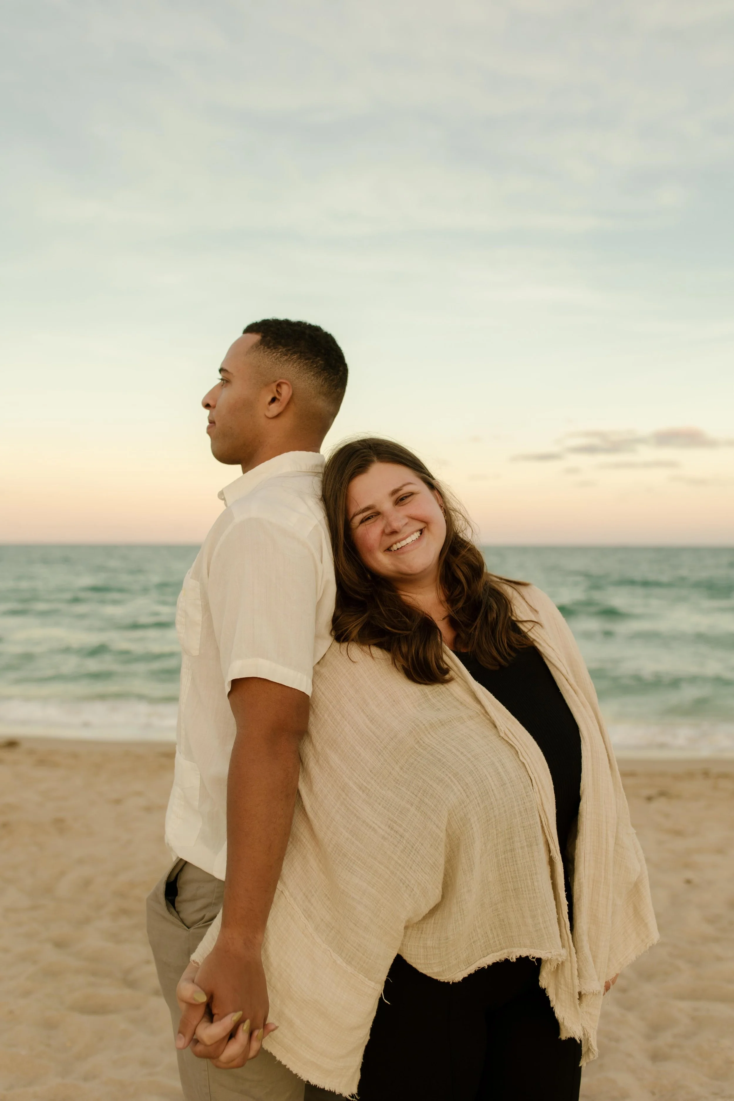 Couple standing back-to-back holding hands on the shoreline, soft pastel sky and ocean behind them, grounded and playful all at once