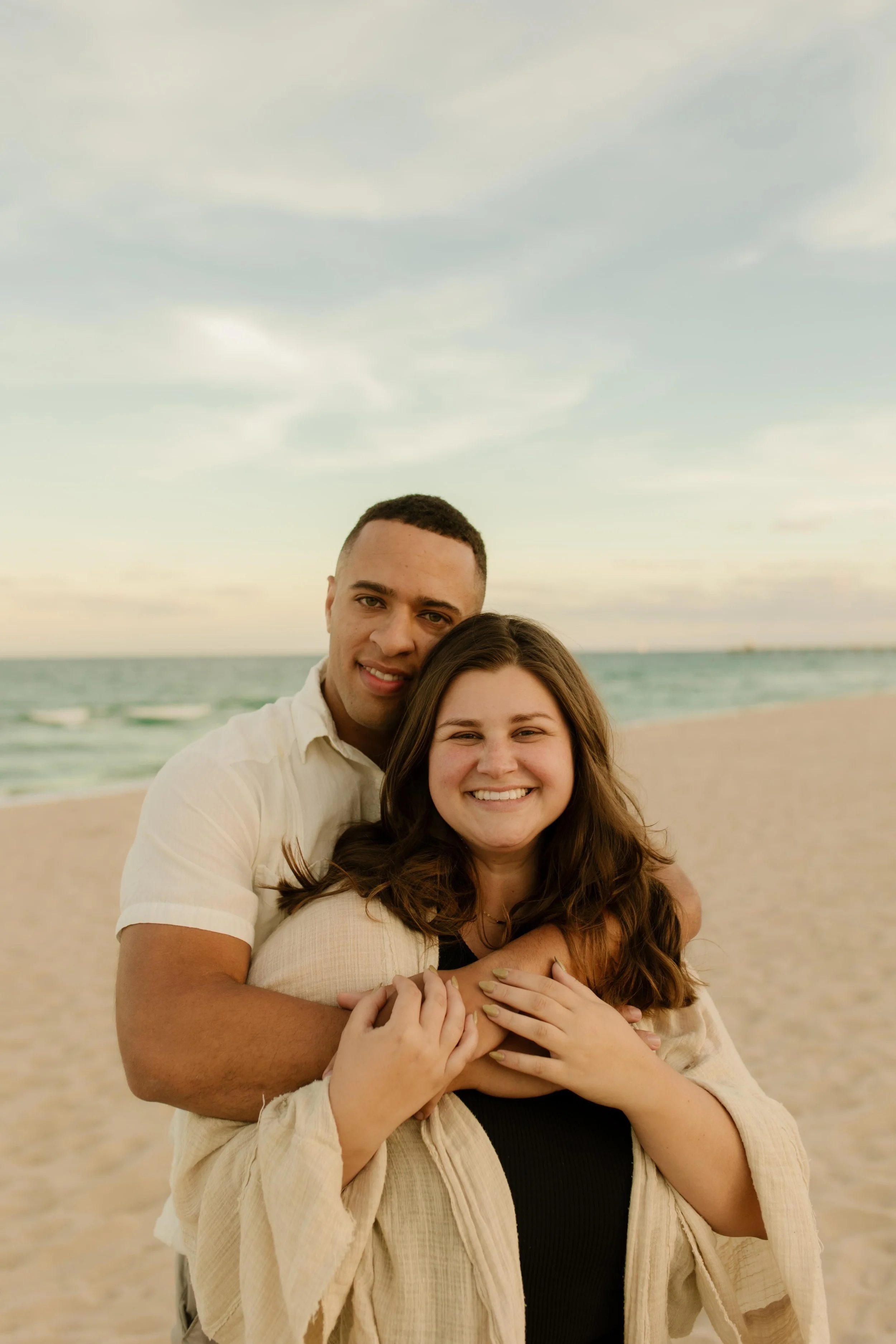 Couple standing on the beach at sunset, arms wrapped around each other from behind as they smile toward the camera, a just-engaged glow