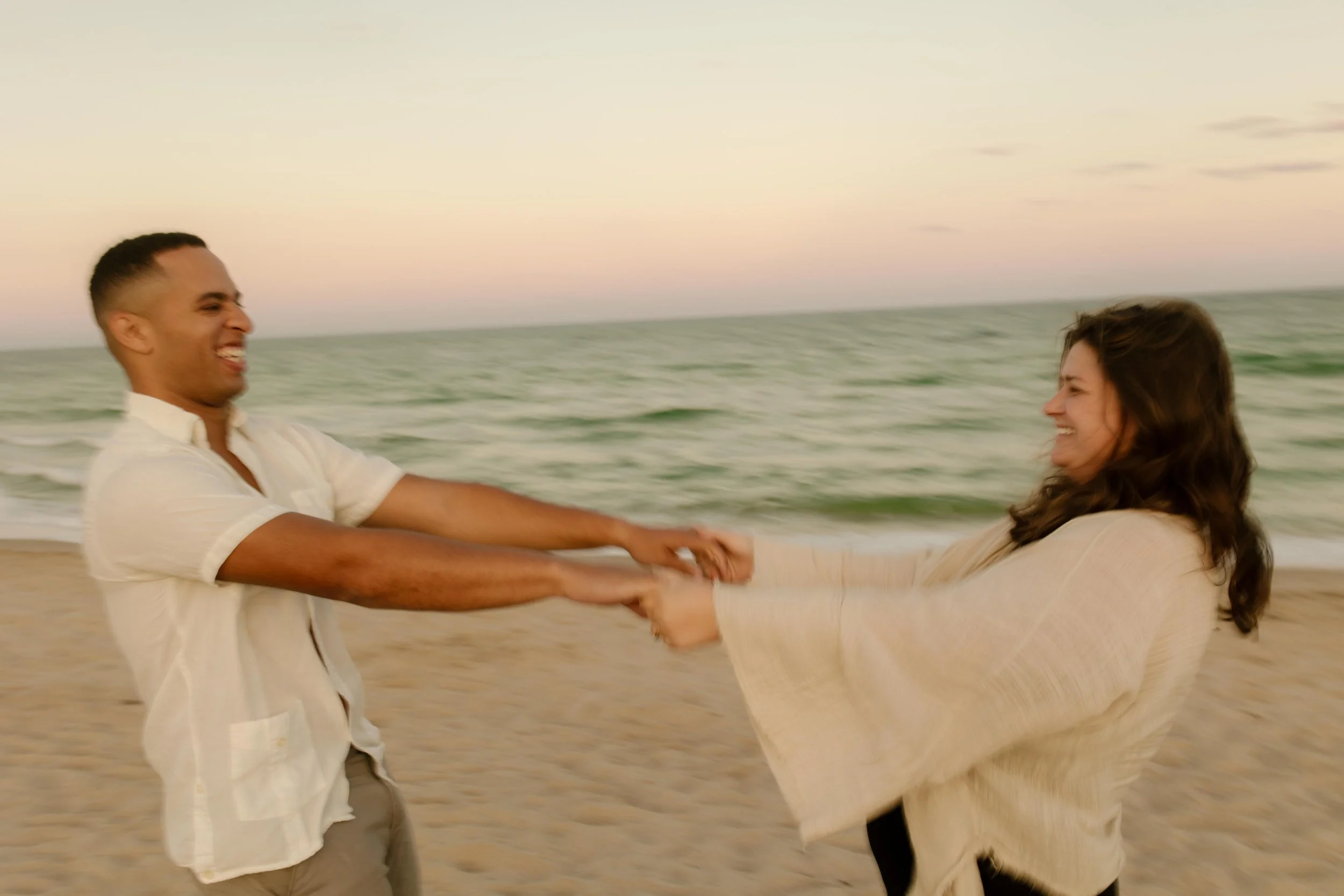 Couple spinning each other around on the shoreline, laughing with arms stretched out and motion blur capturing the joy of just getting engaged