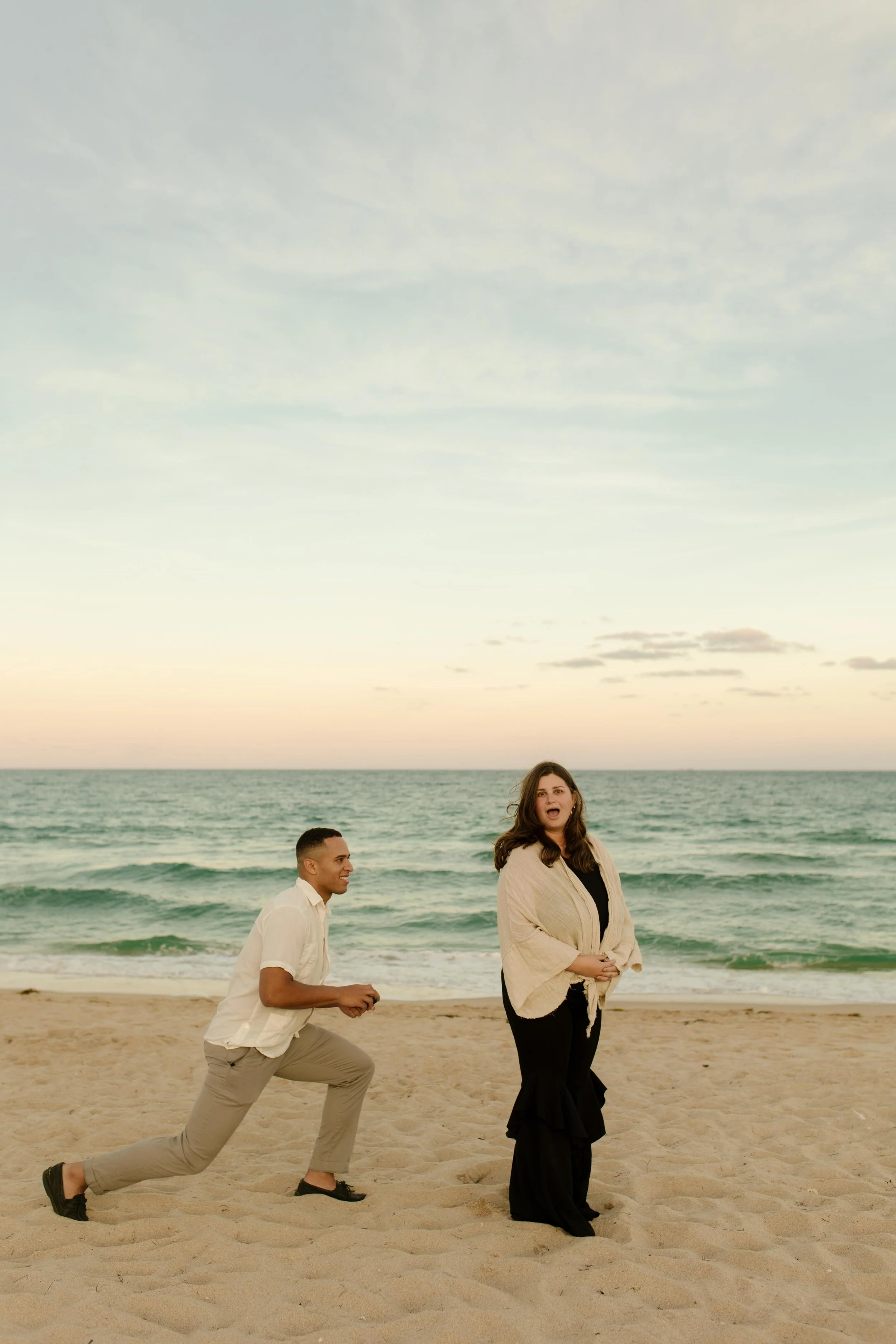 Man down on one knee holding a ring box open as his partner stands in front of him with her hand over her mouth