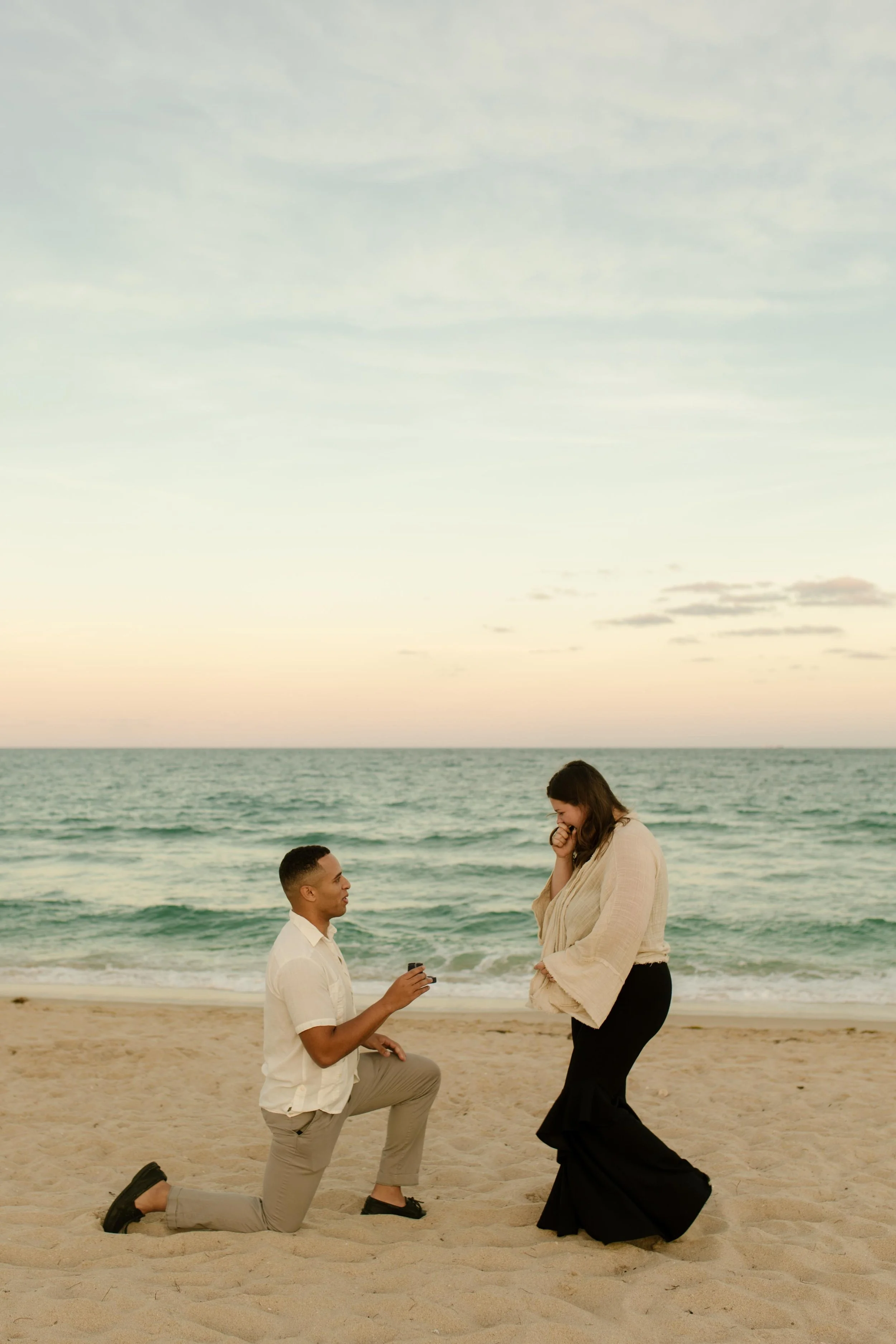 Man kneeling in the sand mid-proposal while his partner turns toward him in shock, ocean waves rolling behind them in soft pastel light