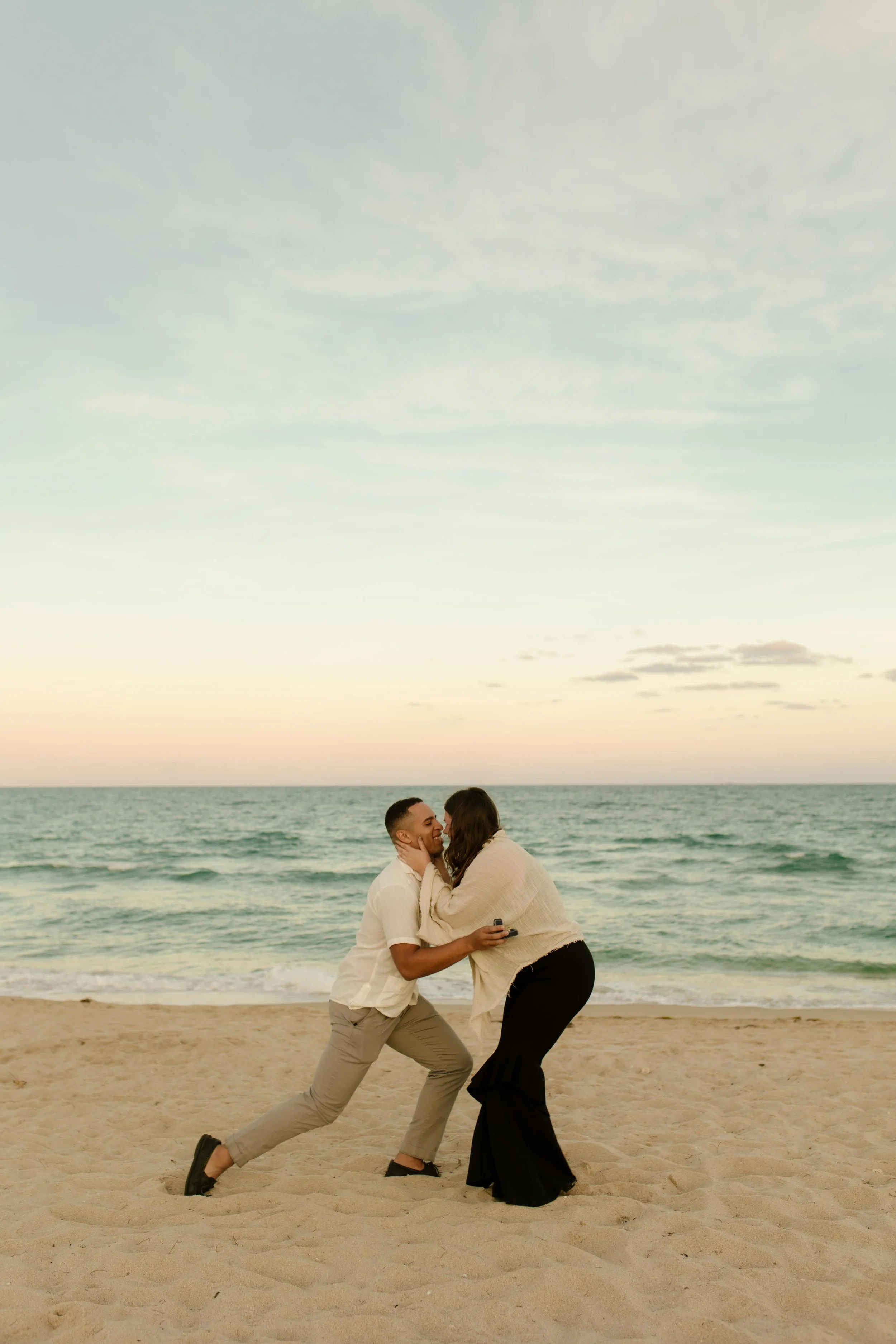 Man down on one knee on a sandy beach holding his partner’s face as she leans in, overwhelmed and emotional in a scene that captures how to plan a surprise proposal that feels intimate and real