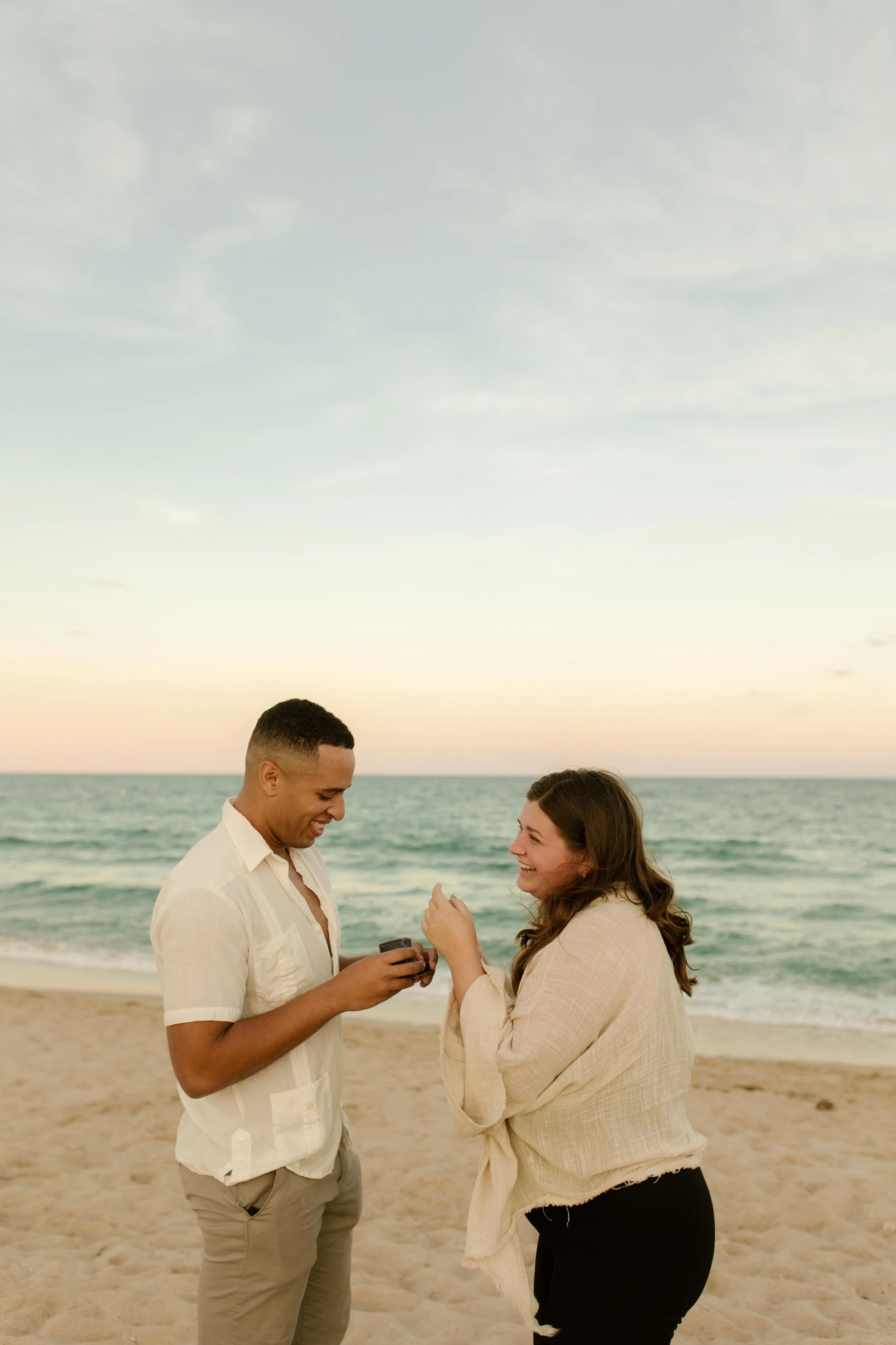 Man holding an open ring box while smiling at his partner as she reacts with laughter and disbelief on the sandy beach, a candid moment showing how to plan a surprise proposal that feels genuine