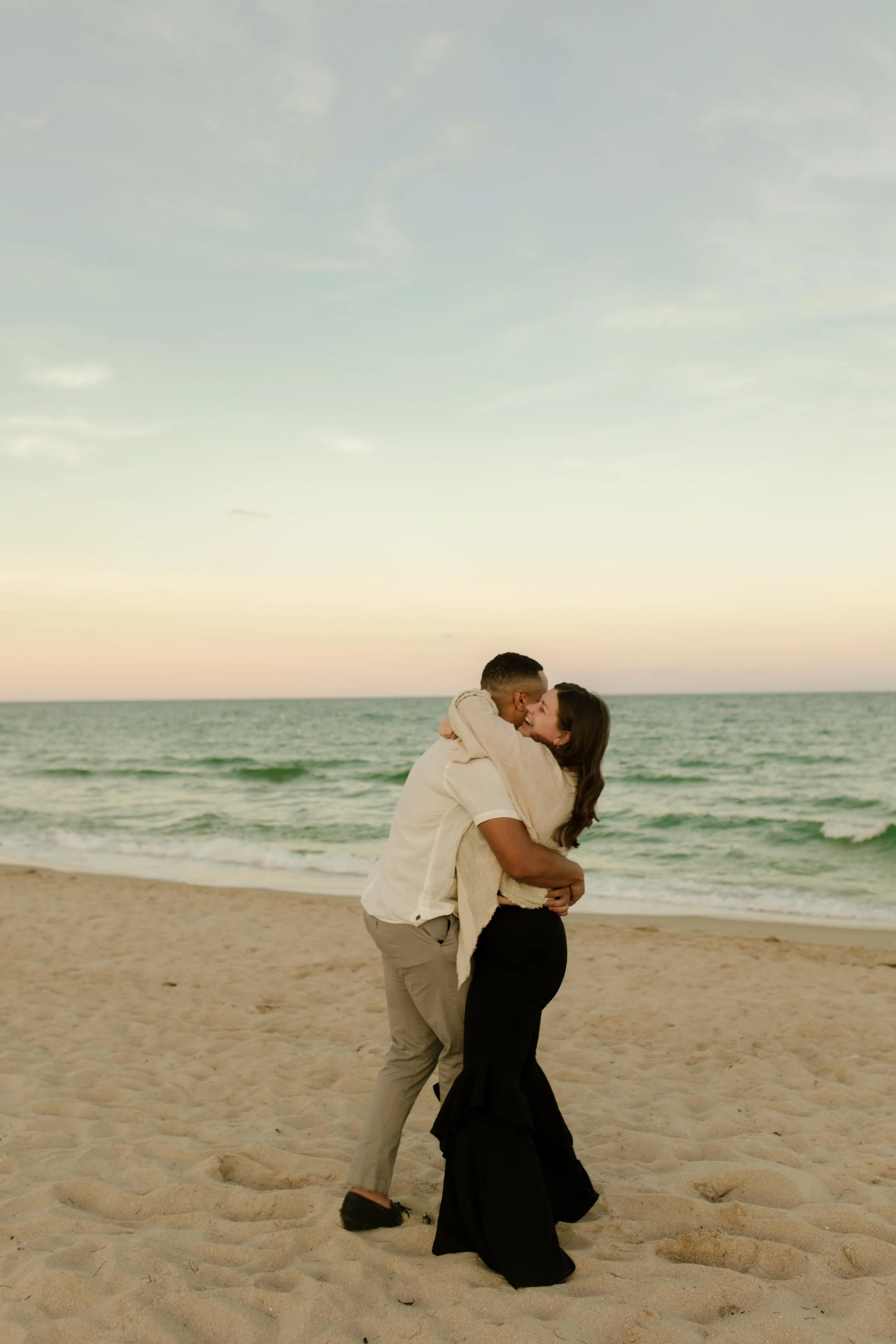 Couple wrapped in a tight embrace on the shoreline, kissing as the sky fades into soft pastels behind them