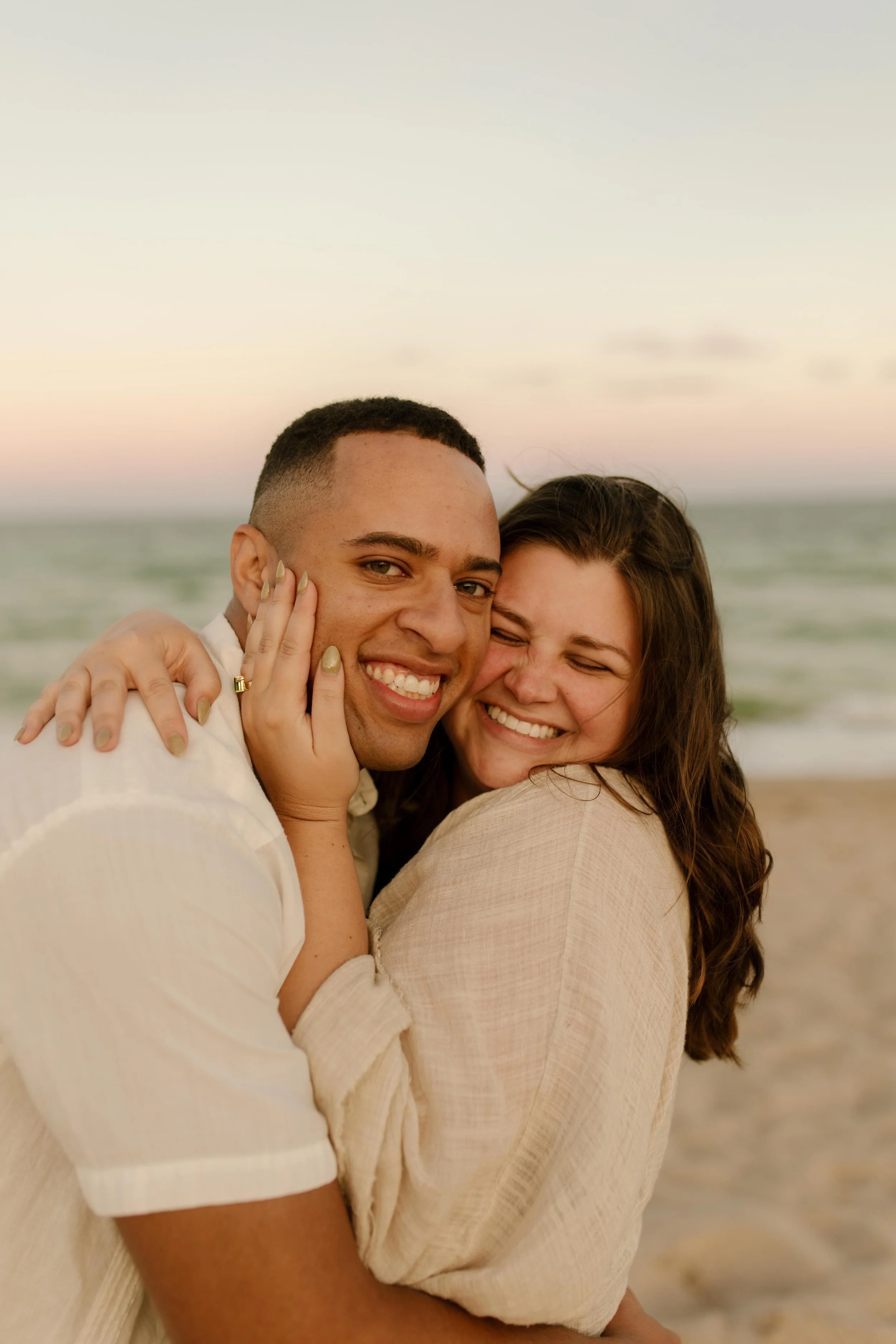 Close-up of a couple hugging tightly on the beach, her hand on his cheek and engagement ring visible, pure joy radiating from both of them