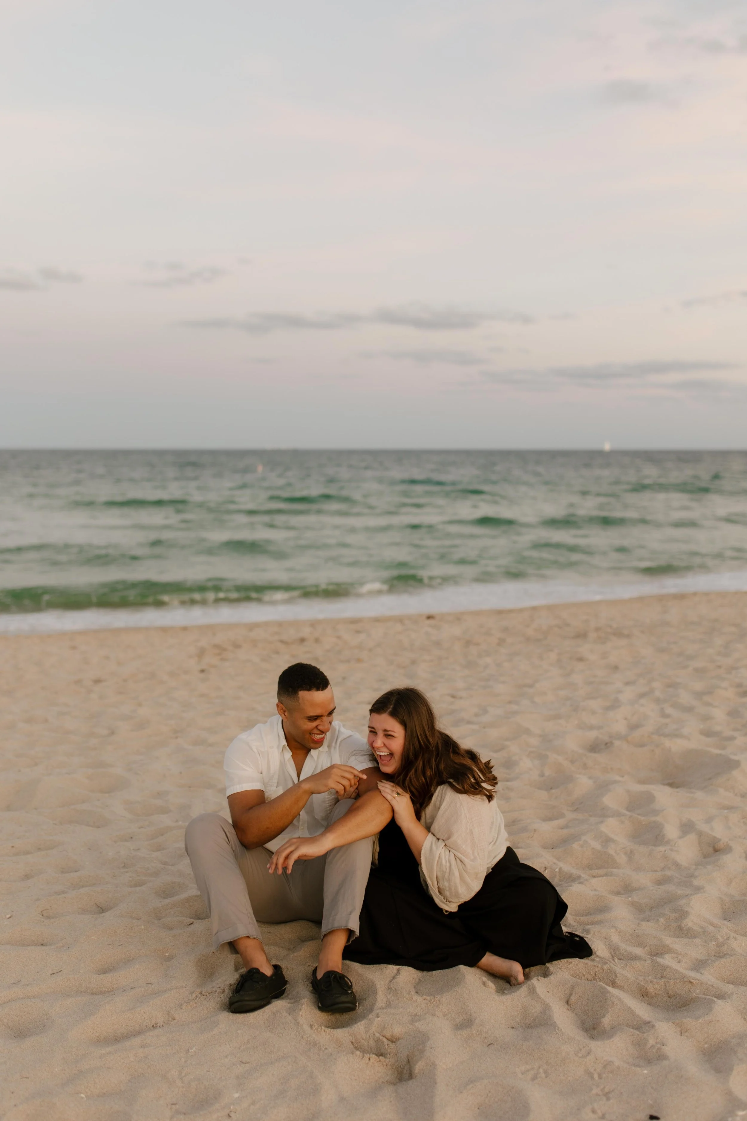 Couple sitting on the sand laughing together, bodies turned inward and completely lost in their own little world after the proposal
