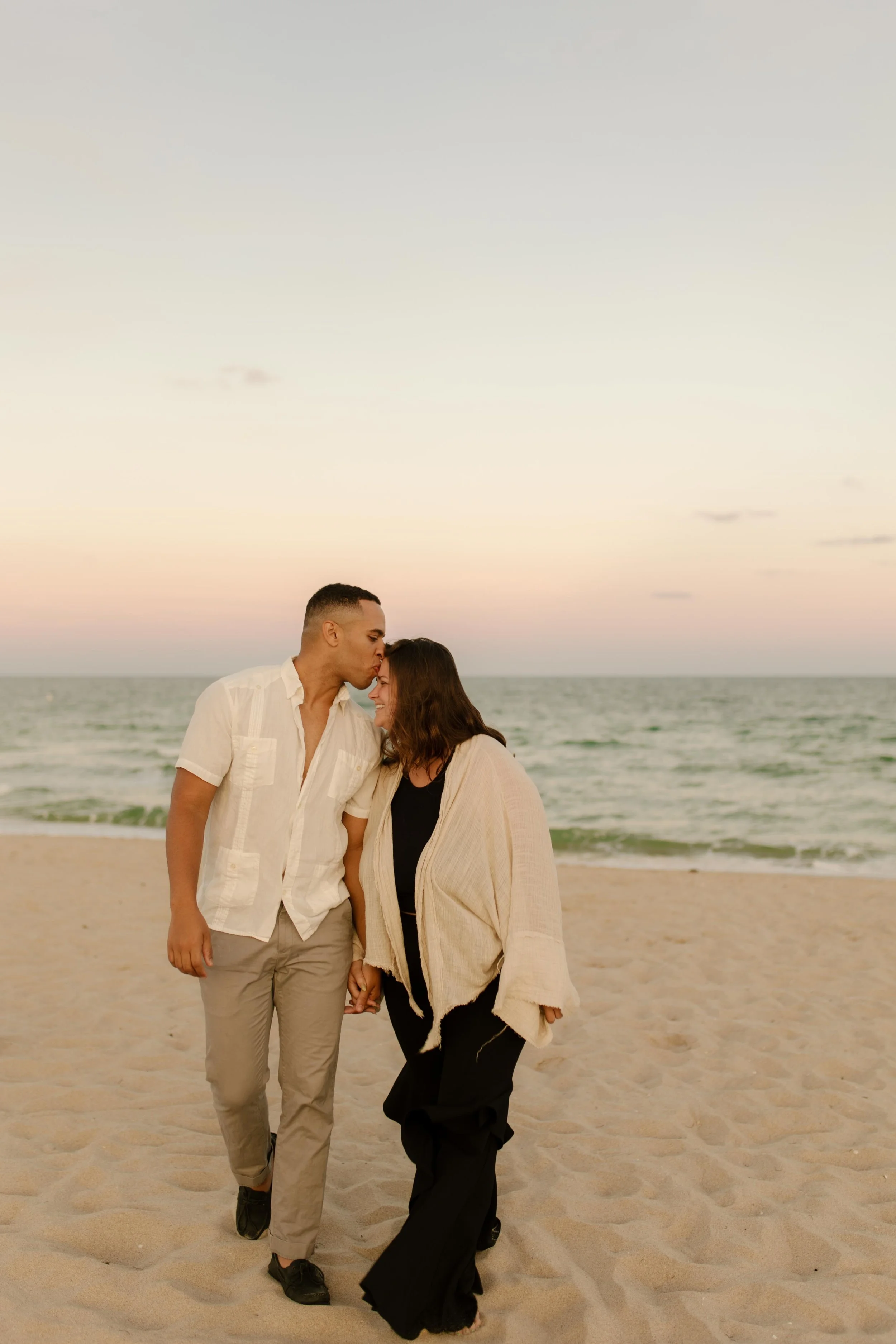 Couple walking hand in hand along the shoreline at sunset, foreheads touching as they move together in a quiet just-engaged haze