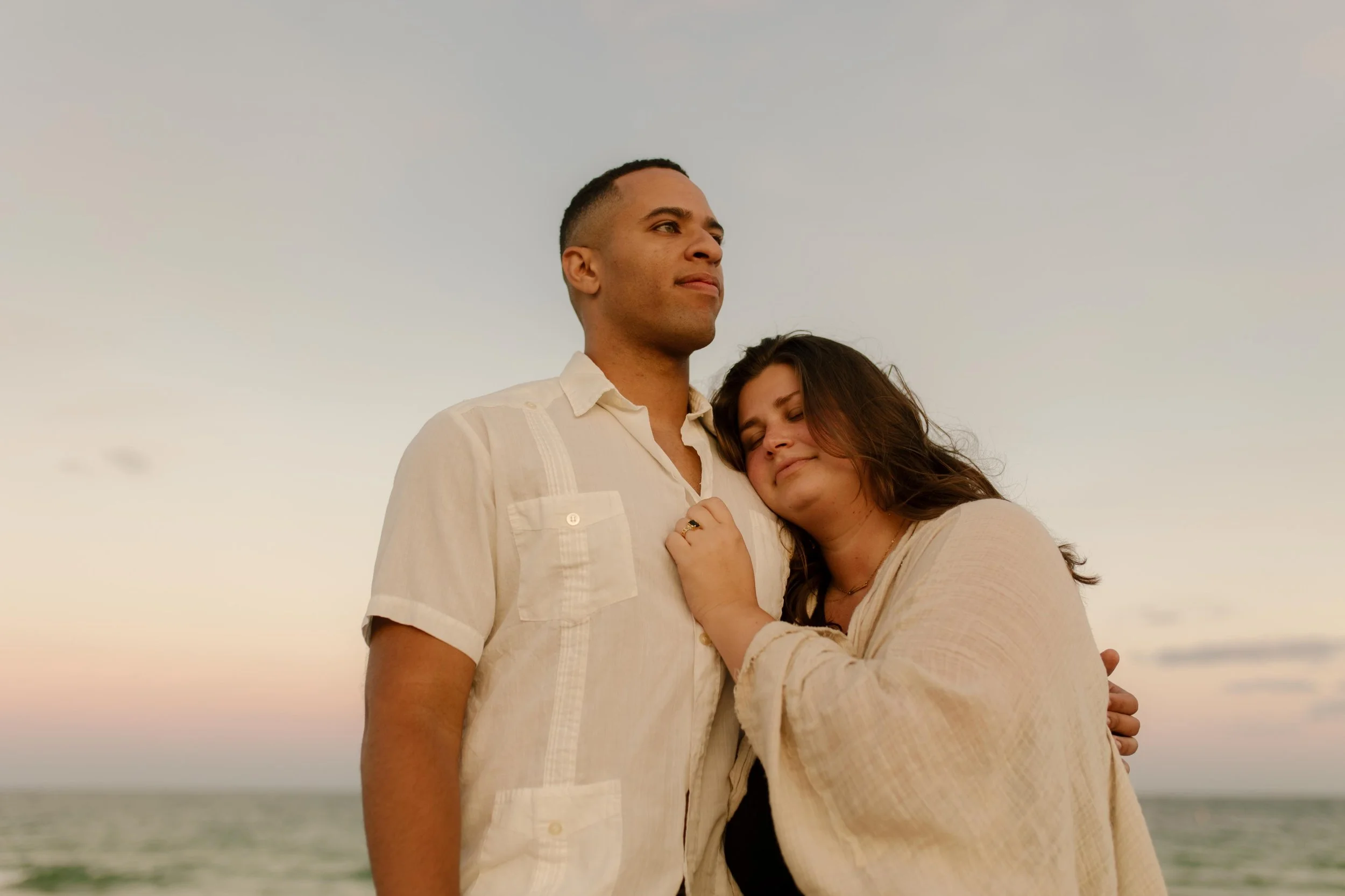 Couple standing together on the beach in a quiet, reflective moment, her head resting on his shoulder as the ocean stretches out behind them