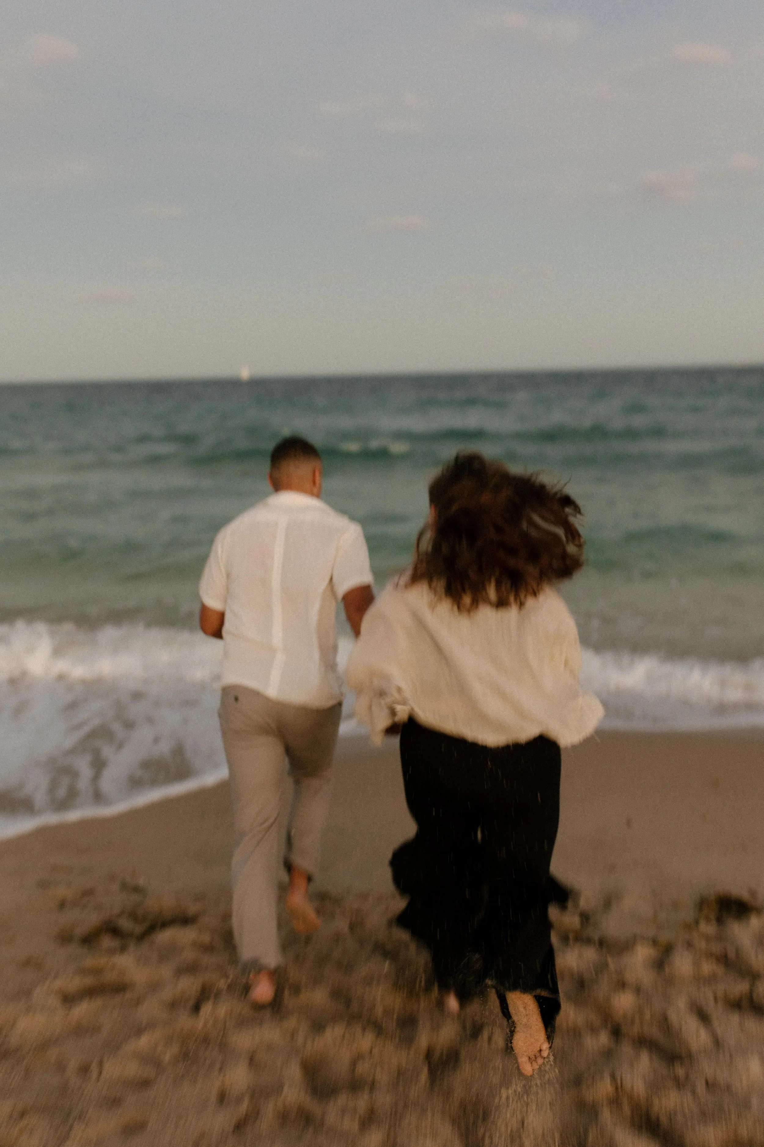 Couple running hand in hand toward the ocean, barefoot and carefree, sand kicking up behind them in a spontaneous celebration after getting engaged