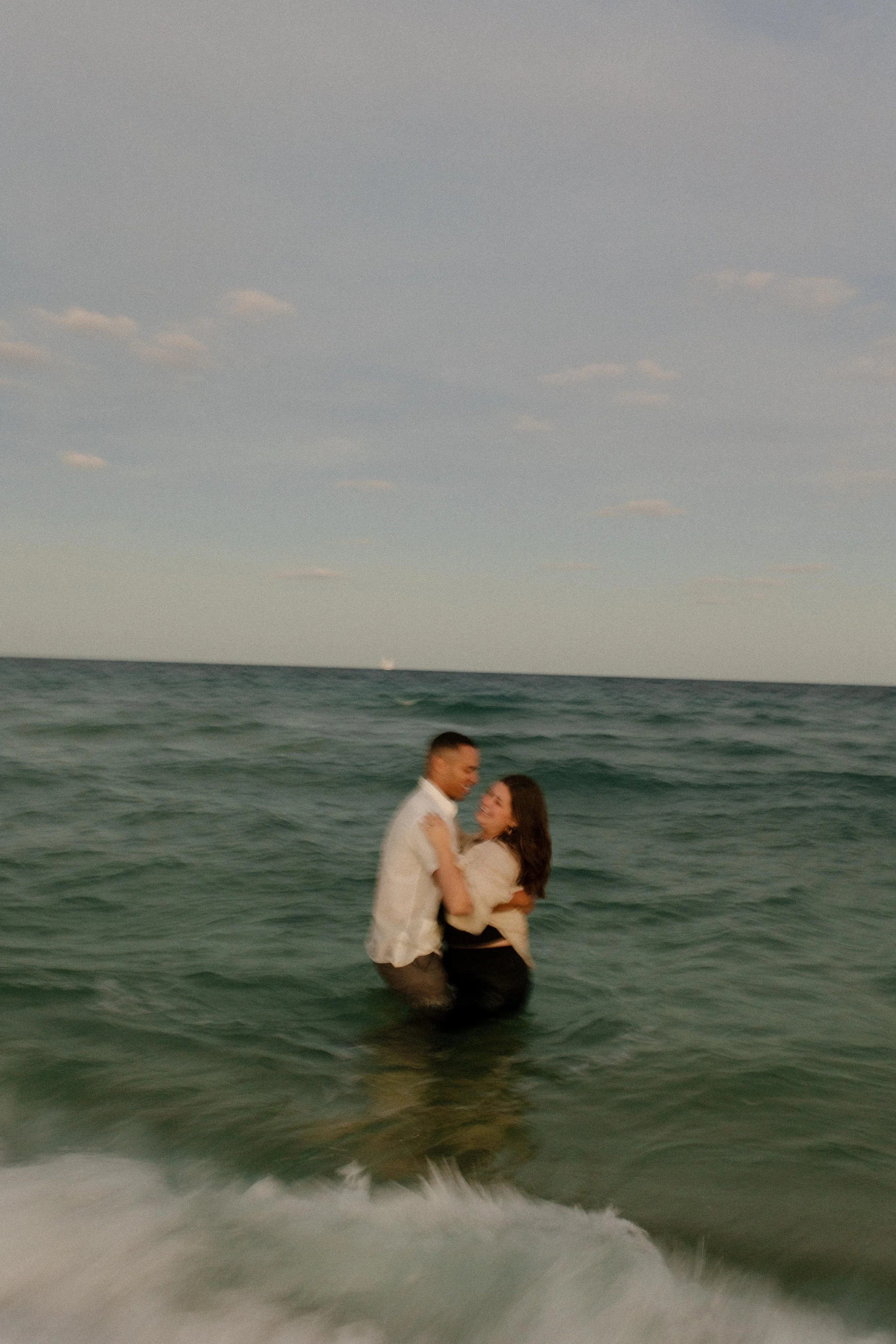 Couple standing waist-deep in the ocean holding each other as waves move around them, a playful and unfiltered moment