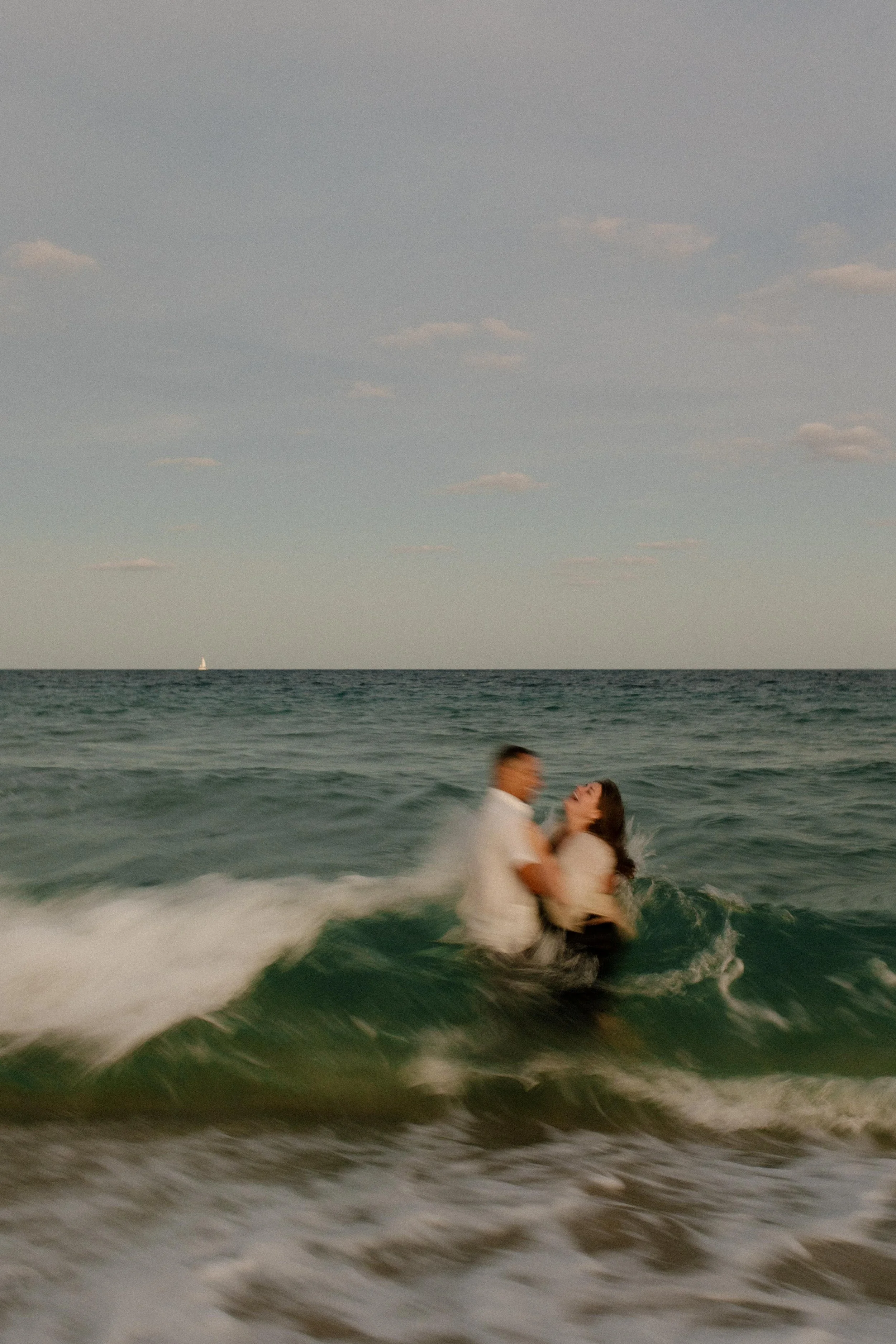 Couple laughing as a wave crashes into them while they hold each other in the water, motion blur capturing the chaos and fun of the moment