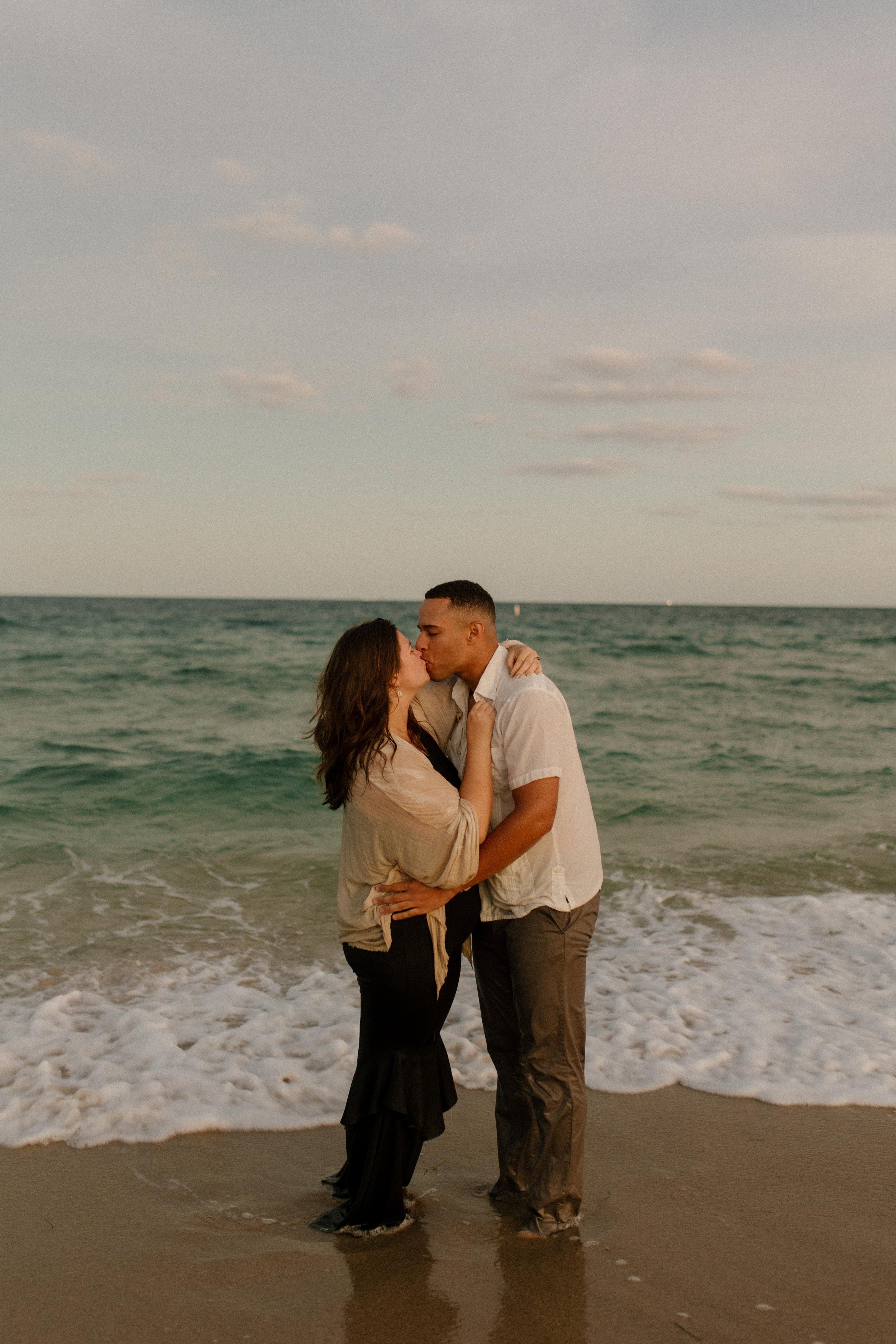 Couple kissing at the shoreline with waves washing over their feet, fully present in the just-engaged feeling