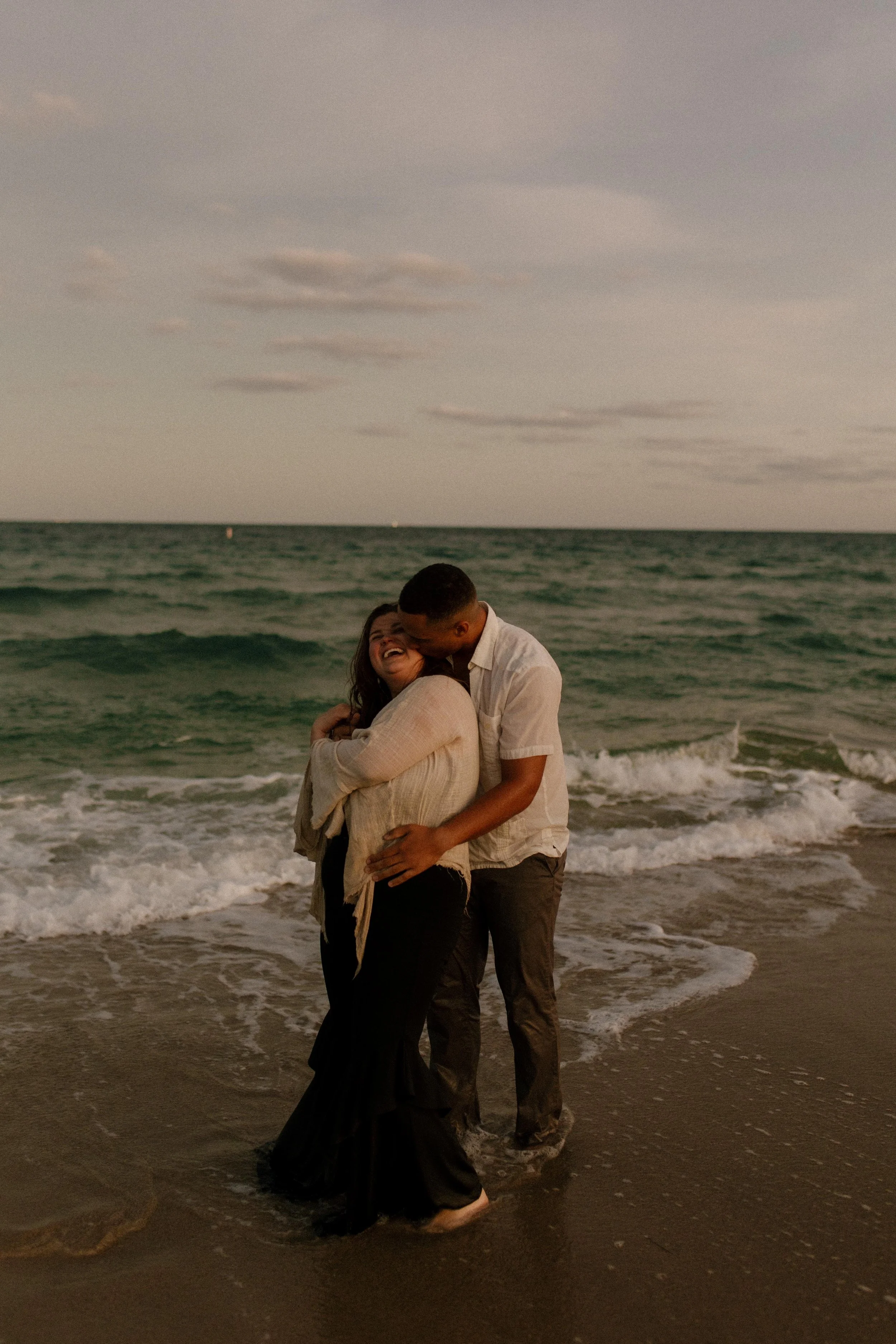 Couple standing at the edge of the ocean as waves crash around them, laughing and holding each other in golden light after a beach proposal