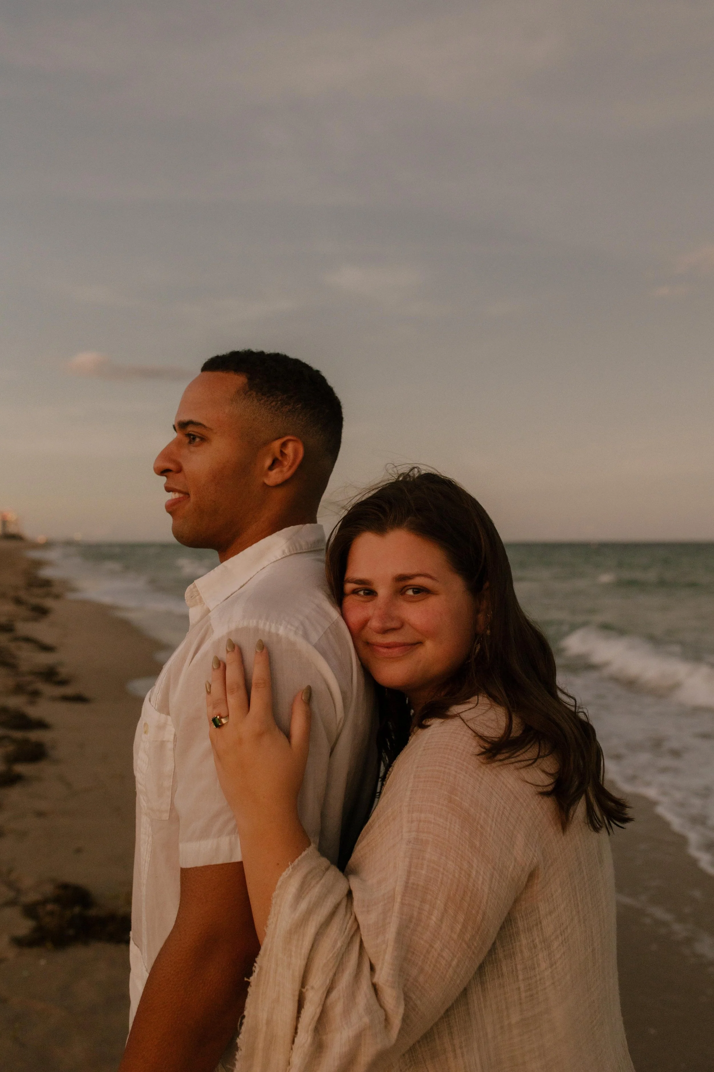 Woman hugging her partner from behind on the beach, smiling softly toward the camera with her hand resting on his shoulder and engagement ring visible