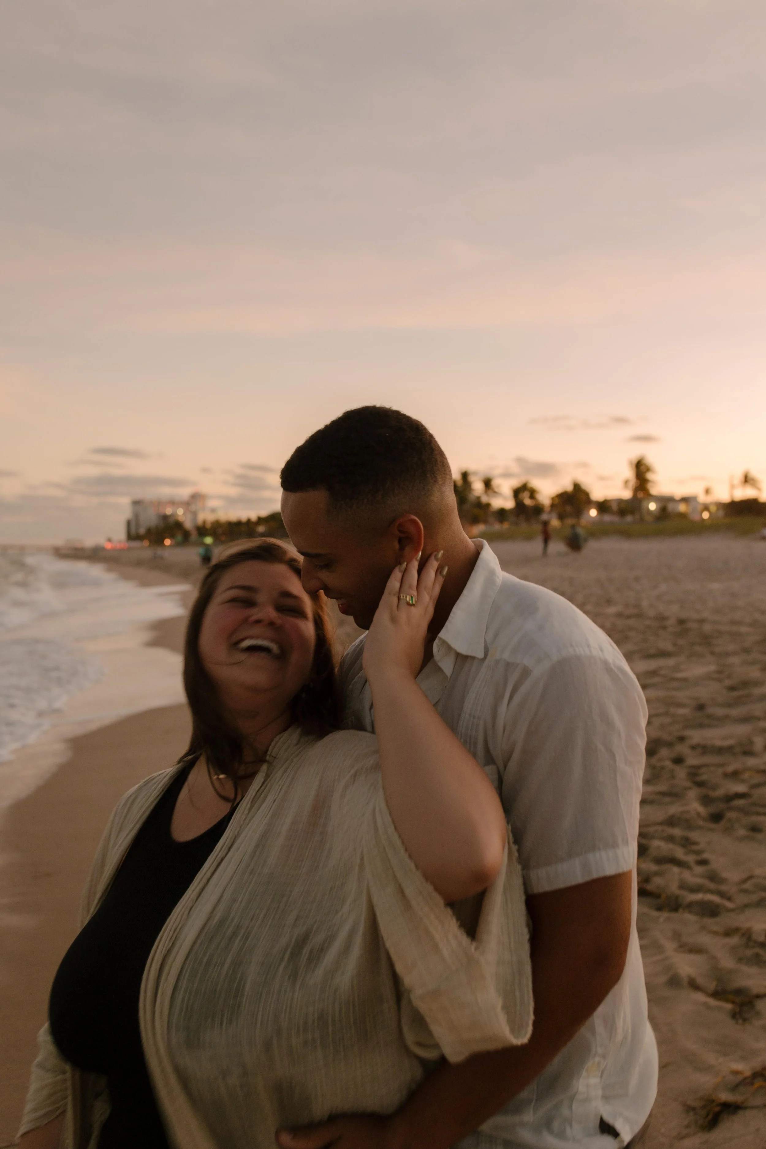 Couple standing close together on the beach at sunset, laughing as she touches his face and her engagement ring catches the light