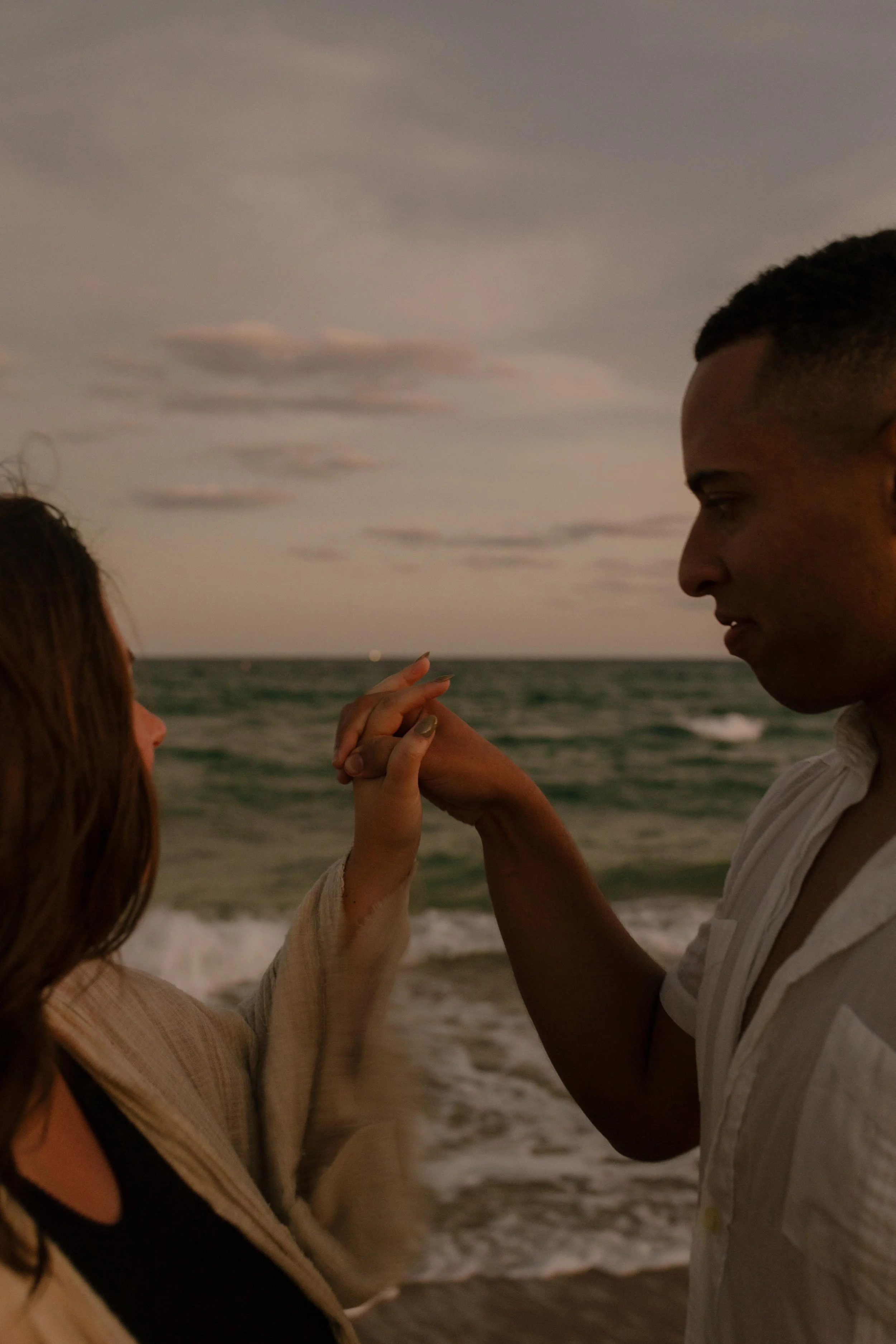 Close-up of a couple holding hands near the shoreline, her engagement ring visible as he lifts her hand gently in a quiet, intimate moment