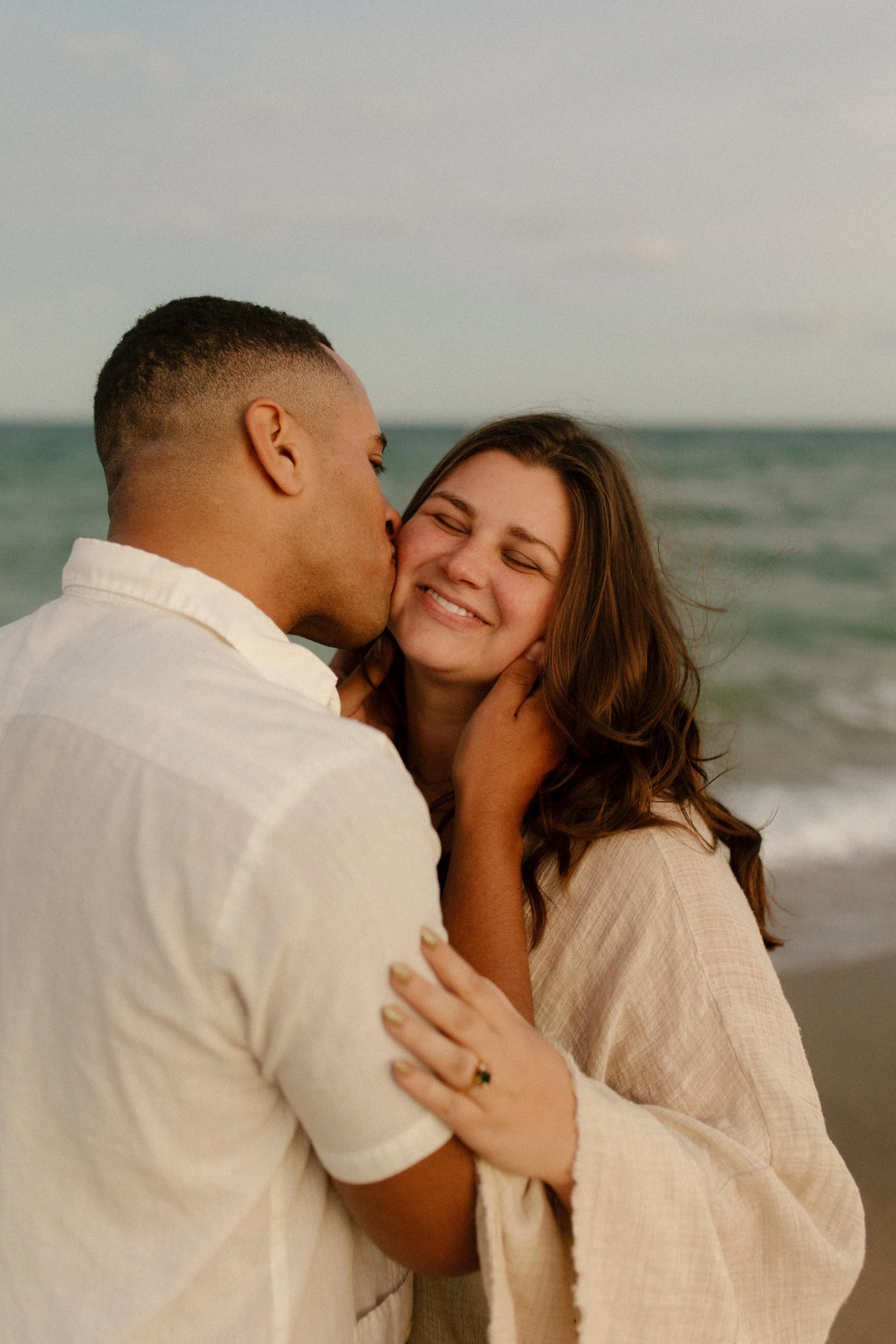 Man kissing his partner’s cheek as she smiles with her eyes closed, ocean behind them and wind moving through her hair