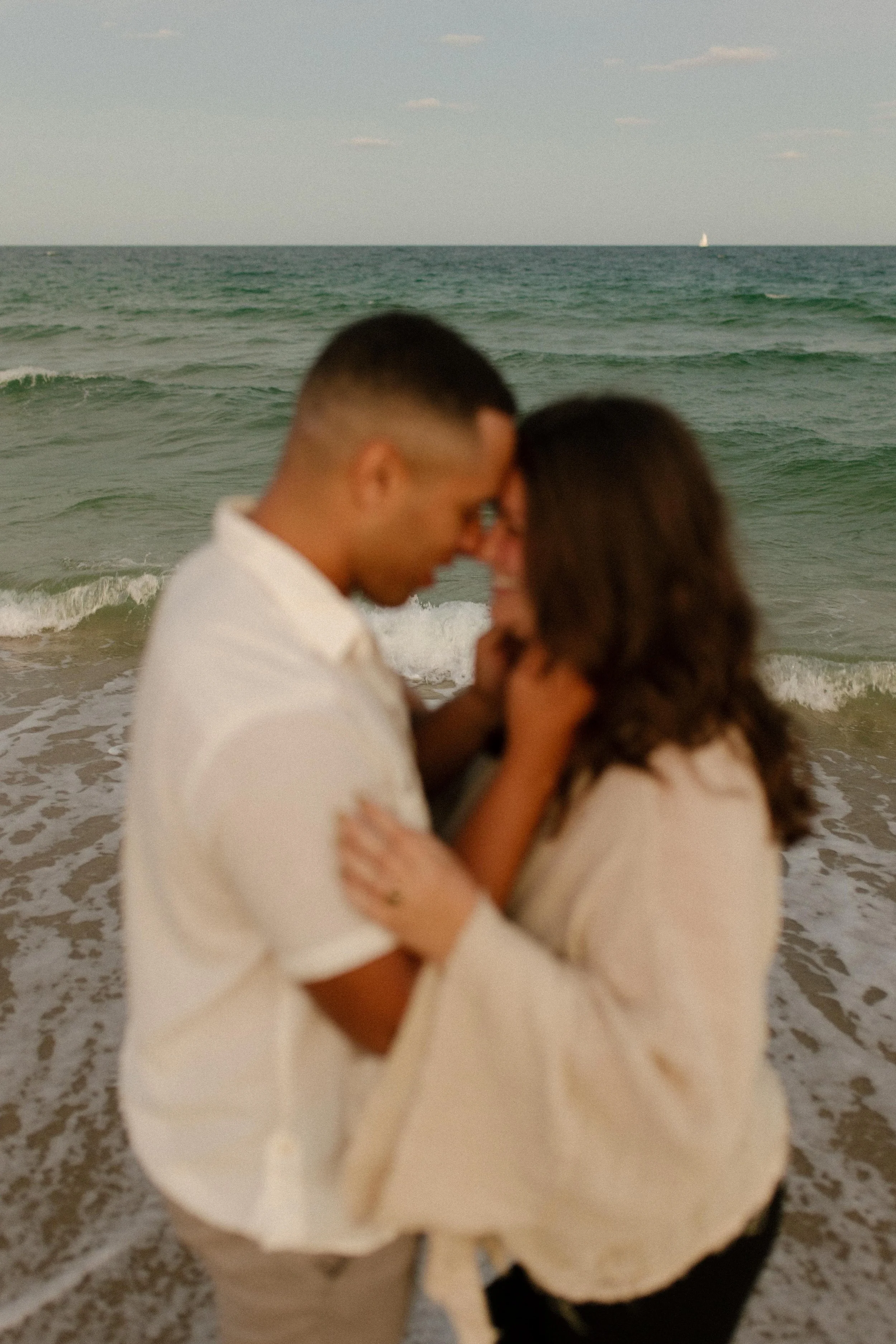 Soft, slightly blurred image of a couple leaning their foreheads together at the water’s edge, capturing the emotional haze right after getting engaged