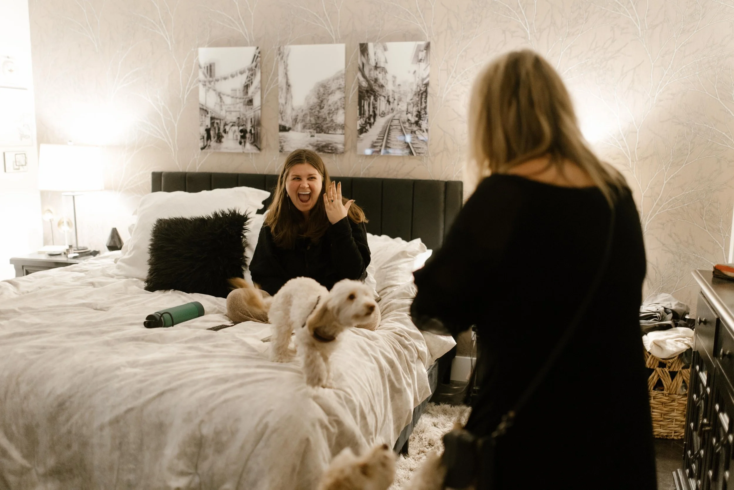 Woman sitting on a bed holding up her hand to show her engagement ring while smiling widely, two small dogs jumping around her in excitement