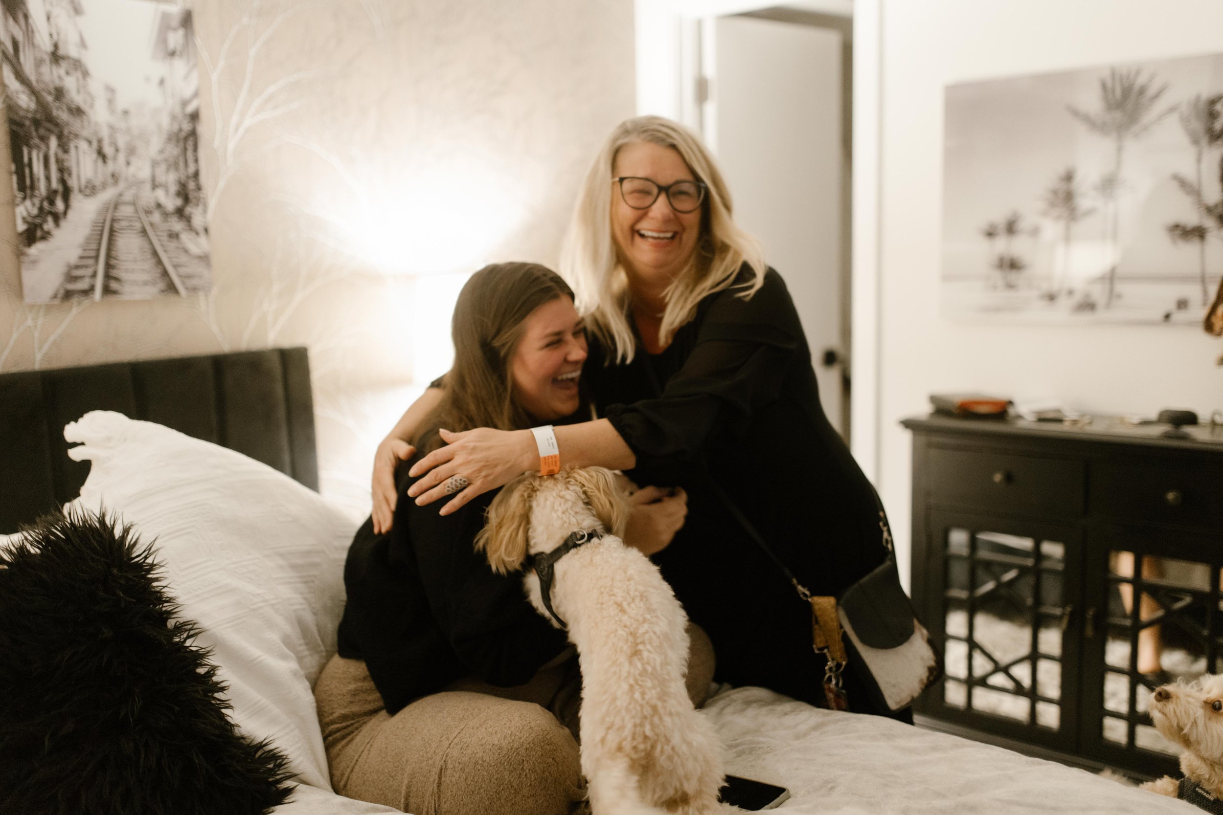 Woman hugging an older woman tightly on a bed while laughing, a small dog jumping up between them, celebrating the just-engaged moment
