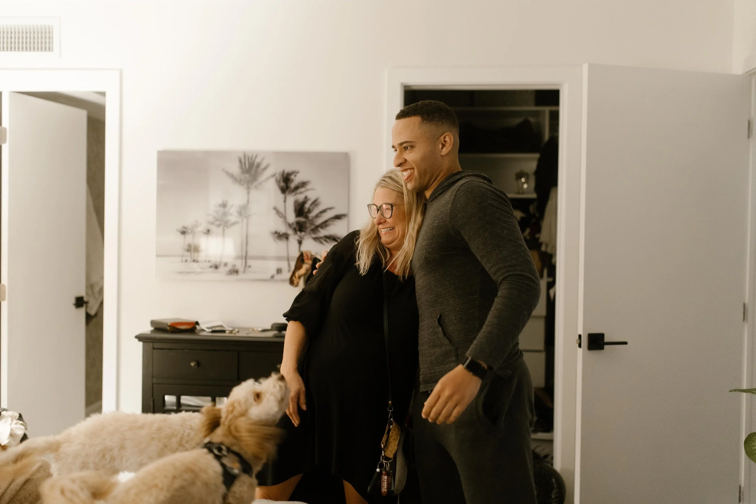 Man hugging a woman in a cozy bedroom while two small dogs jump up excitedly beside them, a candid pre-proposal moment that reflects how to plan a surprise proposal with help from loved ones