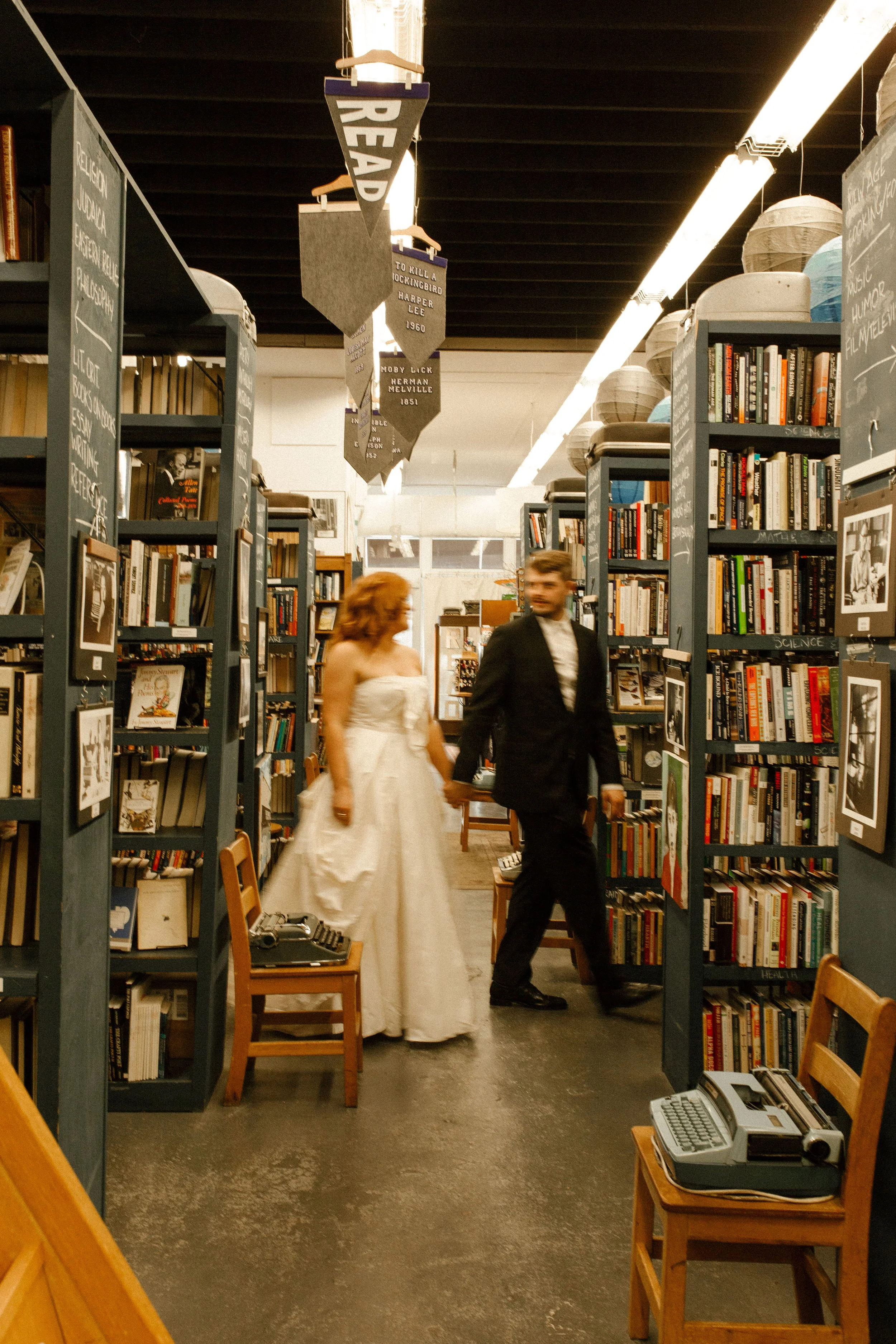 Bride and groom walk hand in hand down a narrow bookstore aisle, smiling at each other between towering shelves of books.