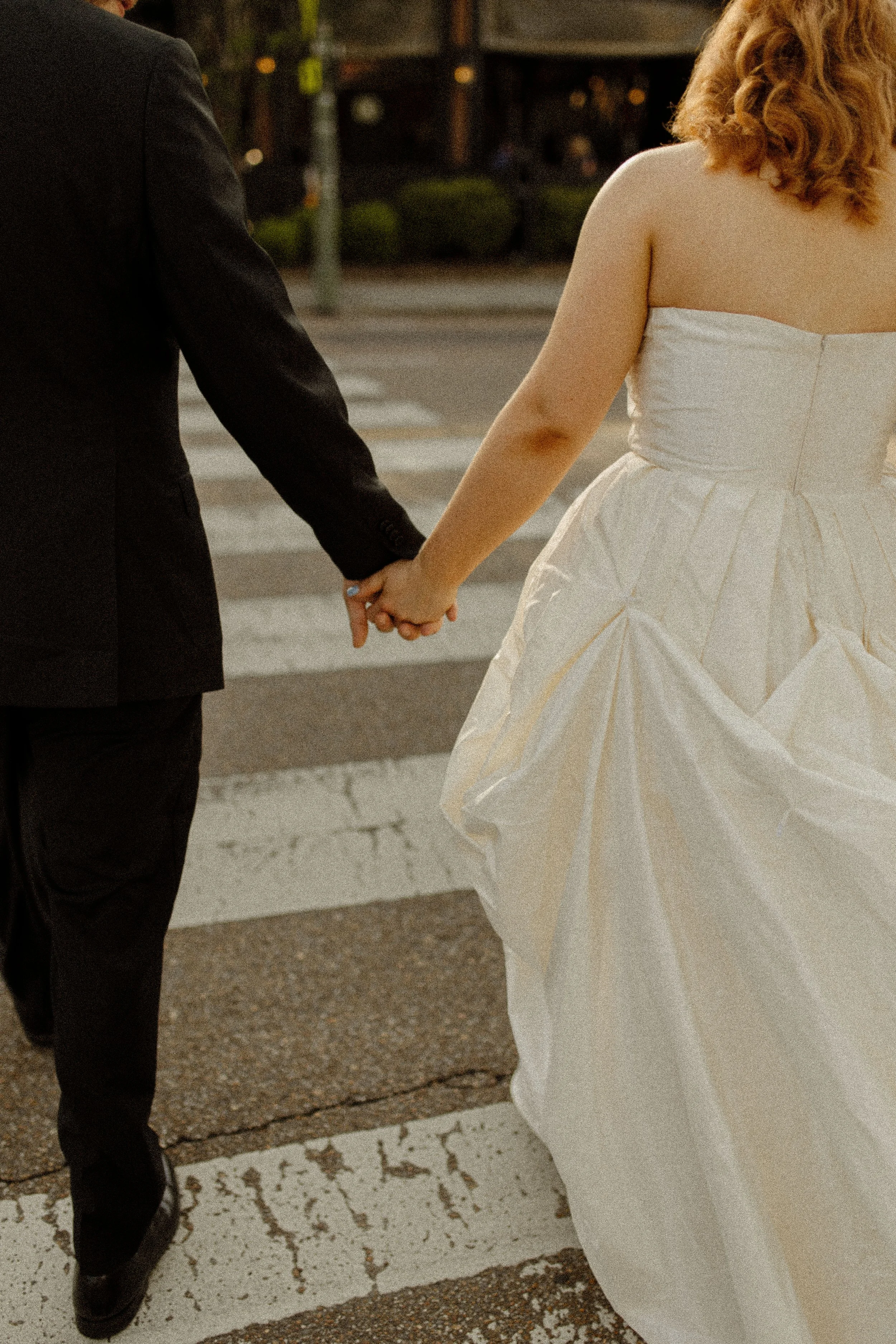 Close-up of a couple holding hands while crossing the street, the bride’s dress flowing softly beside them.