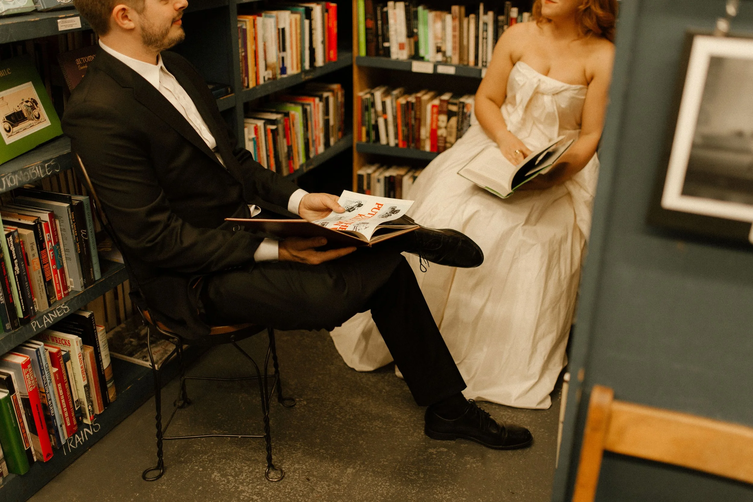 A couple sits across from each other in a cozy bookstore, flipping through books in quiet, intimate pre wedding photos moment surrounded by tall shelves.