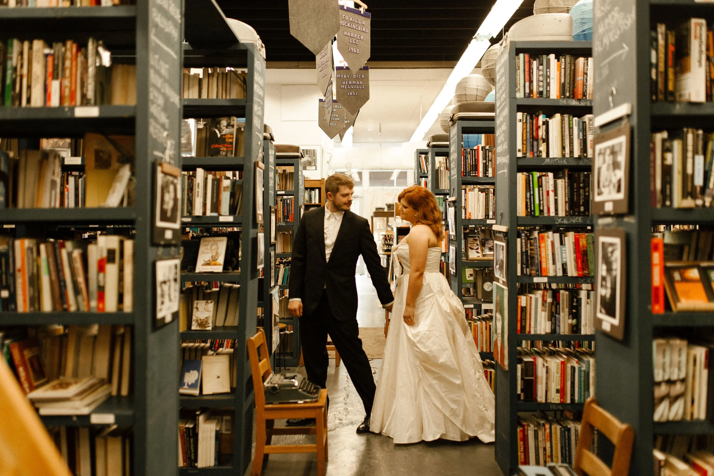 Couple pauses between bookstore shelves, smiling at each other in a quiet, intimate pre wedding photos scene surrounded by books.