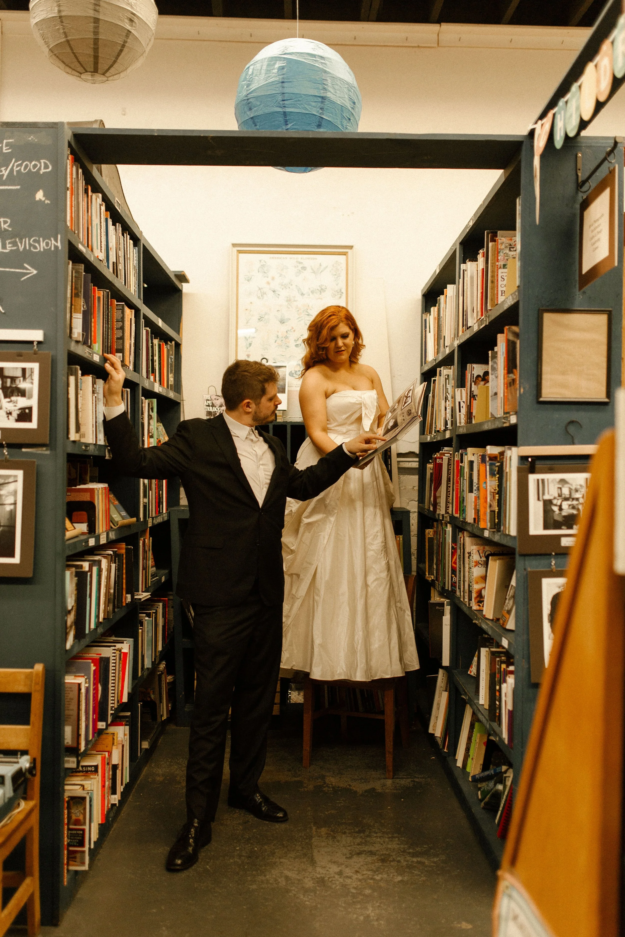 Groom leans casually against a bookshelf while bride stands elevated reading, creating a playful and editorial pre wedding photos scene.