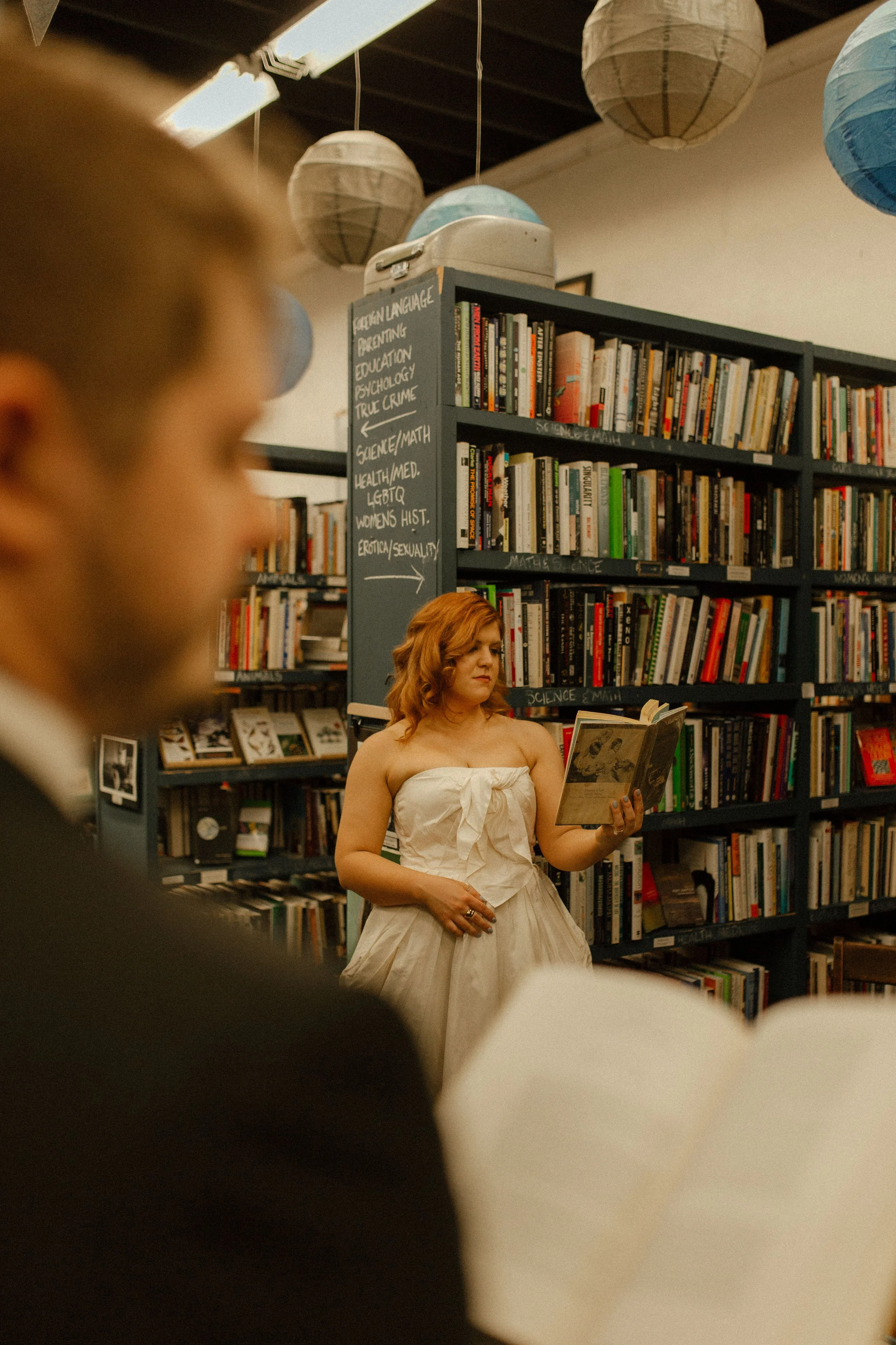 Bride flips through a book while groom is softly out of focus in the foreground, adding depth to this candid pre wedding photos moment.