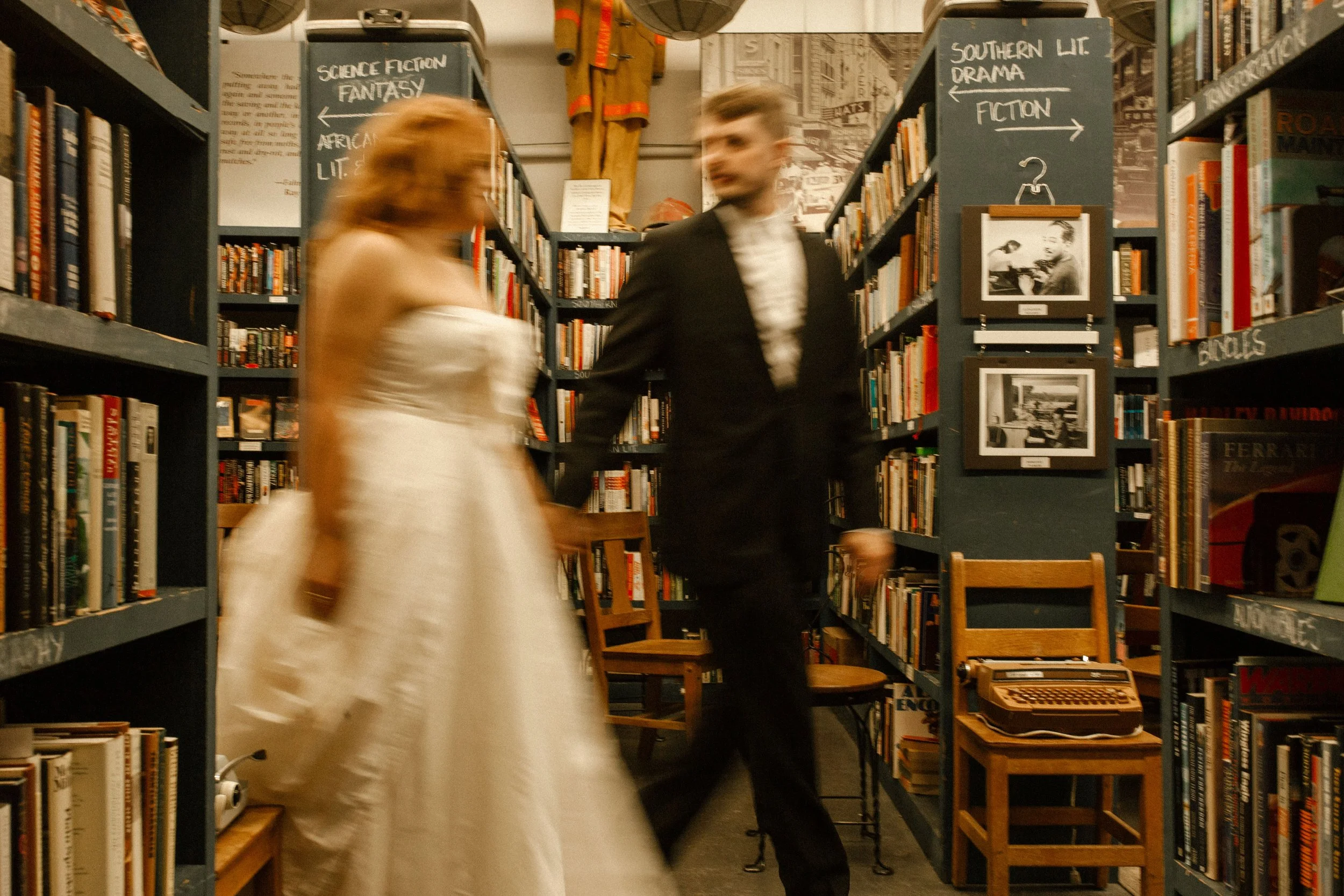 Bride and groom pass each other in a narrow bookstore aisle, motion blurred in a candid pre wedding photos moment between tall shelves.