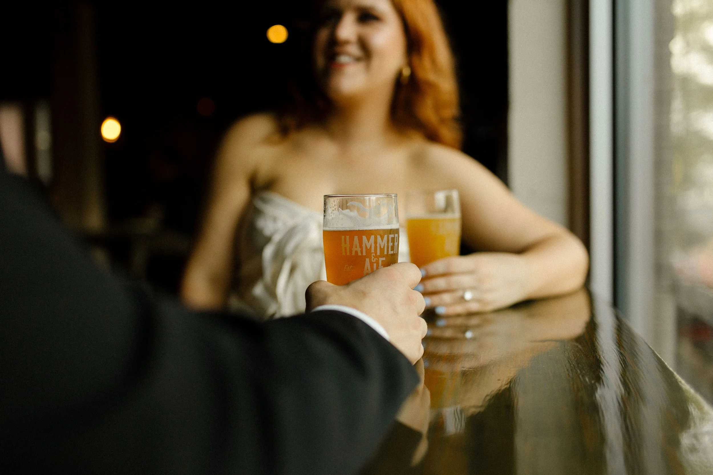 Close-up of hands holding glasses of beer across a bar table, with the bride softly smiling in the background.
