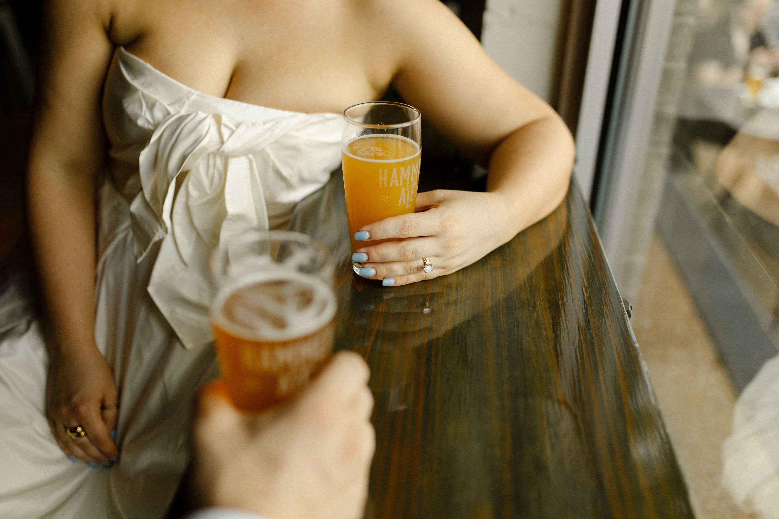 Close-up of bride holding a beer glass at the bar, her engagement ring and soft blue nails visible in a candid detail.
