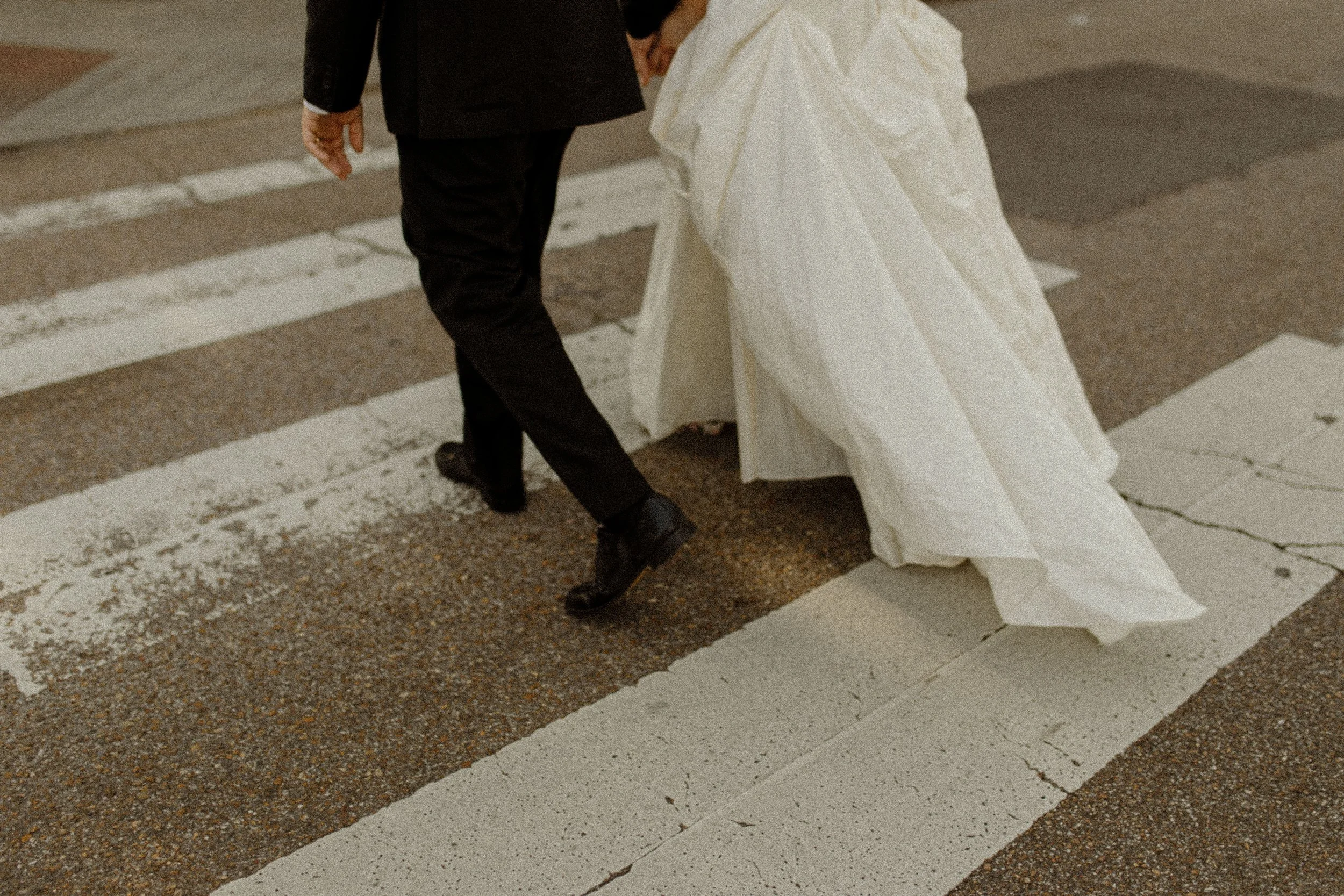 Couple walks across a crosswalk hand in hand, the bride’s dress trailing behind in a candid and romantic pre wedding photos scene.